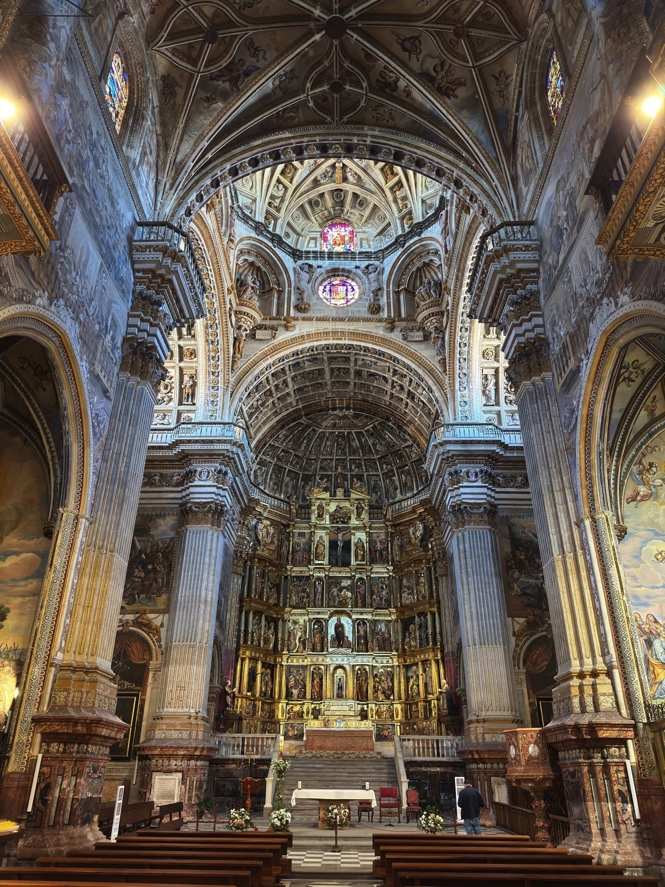 Ornate view of the main altar from the nave, showcasing the intricate gilded altarpiece with multiple tiers of religious figures framed by elaborately carved architecture. The domed ceiling above is richly decorated with painted panels and reliefs, while stained glass windows cast colored light into the grand interior. Towering columns and mural-covered walls add to the sense of grandeur.