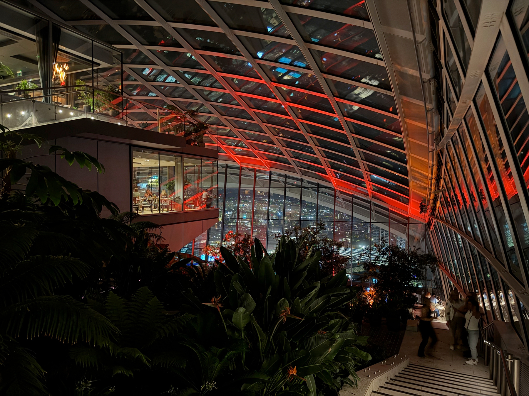 An elevated indoor garden walkway lined with lush plants and a glass roof lit red, where visitors stroll and stop to look out over the city through floor-to-ceiling windows.