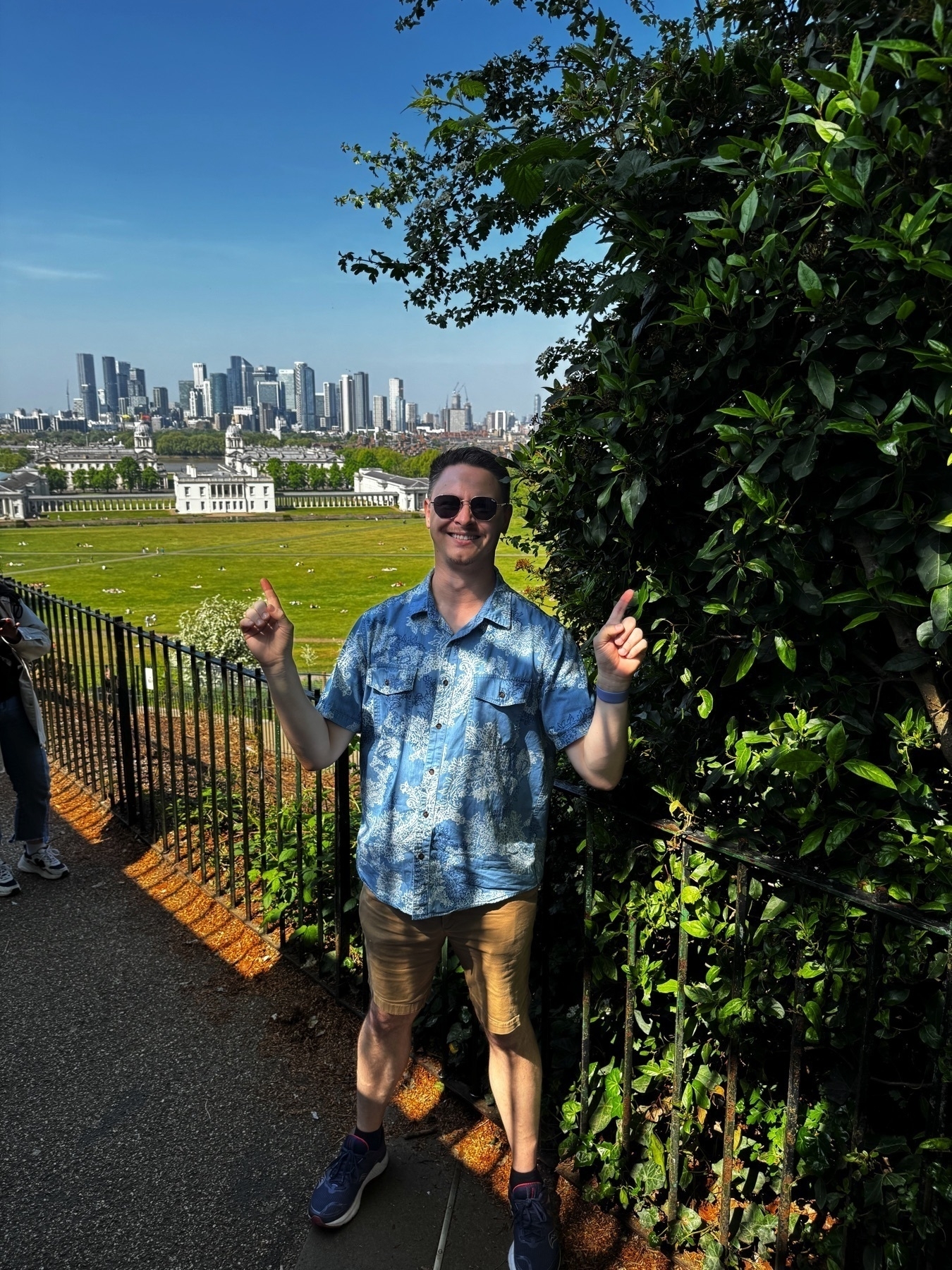 A smiling person poses with both index fingers pointed upward in front of a scenic overlook in Greenwich Park. Behind them, a sweeping view reveals the Queen’s House and Old Royal Naval College with the towering skyline of Canary Wharf in the distance. Trees and manicured lawns fill the middle ground under a cloudless sky.