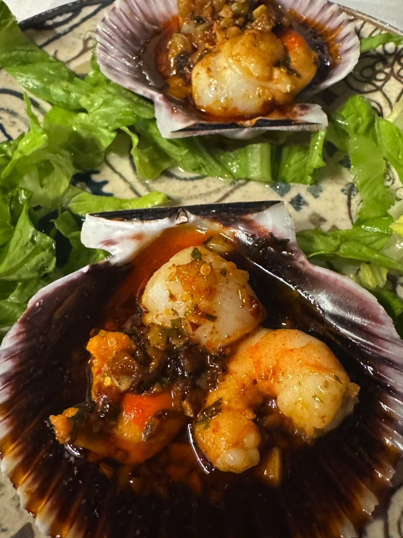 Grilled shellfish served in natural scallop shells, topped with a glistening garlic and parsley sauce that has caramelized slightly at the edges. The scallops appear plump and golden, and they rest on a base of curly lettuce leaves arranged on a decorative plate.