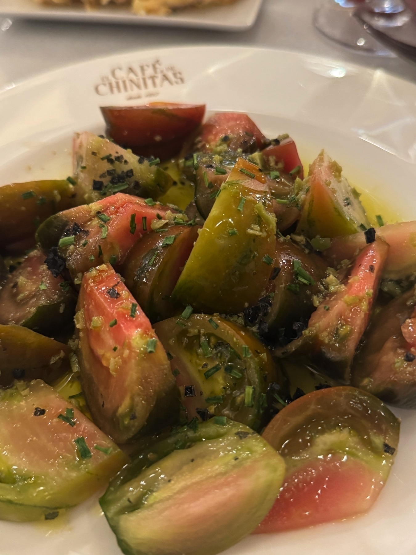 A fresh tomato salad featuring chunks of multicolored heirloom tomatoes dressed in olive oil and sprinkled with flaky salt and chopped herbs. The tomatoes glisten with seasoning, highlighting their ripeness and juiciness.