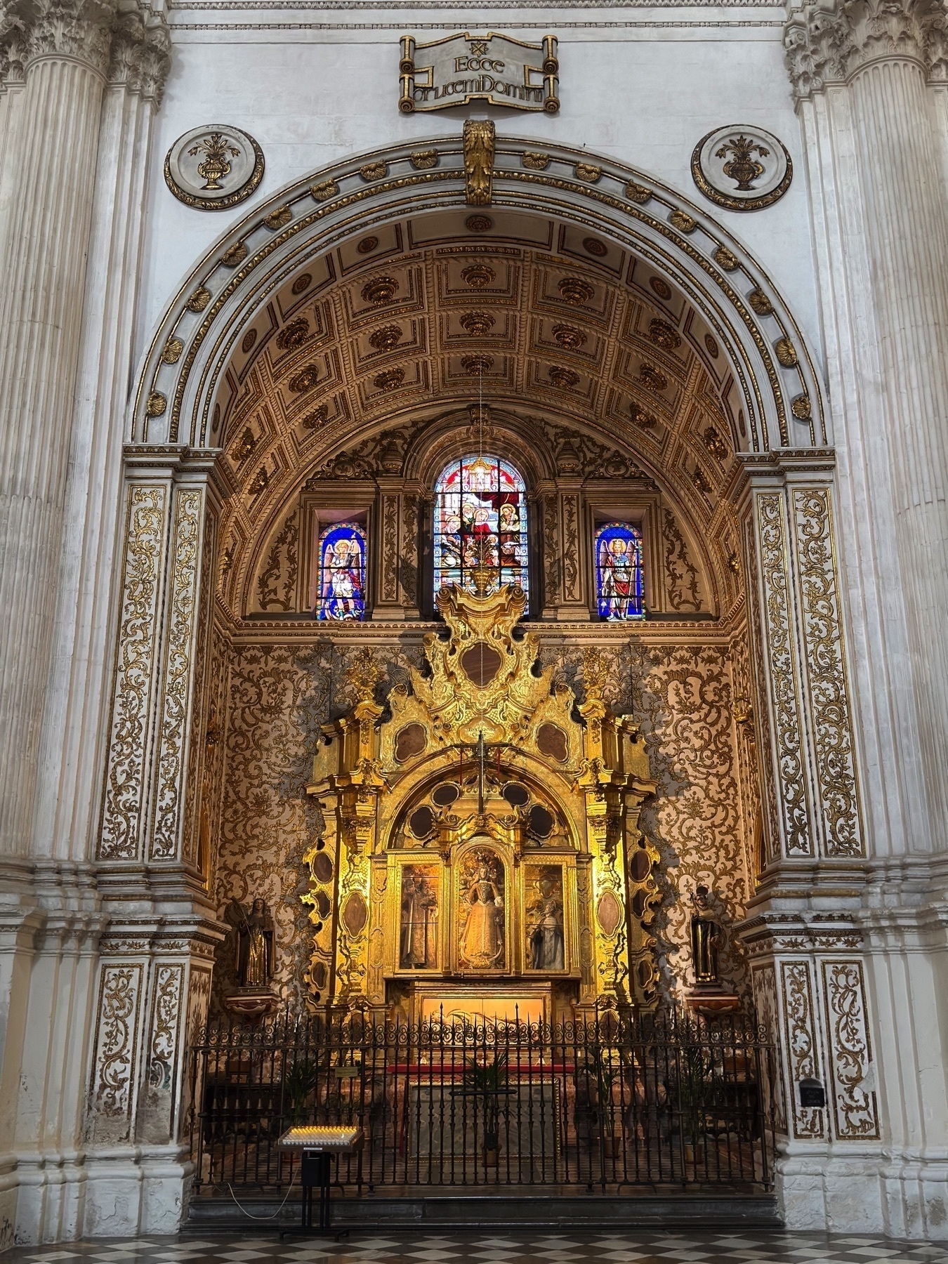 Another elaborately decorated side altar, showcasing a golden Gothic-style altarpiece beneath an arched ceiling richly painted with ornamental patterns. Above the altar, stained-glass windows filter colored light, enhancing the visual warmth of the gold leaf and stone textures. The surrounding area is framed by white marble columns and filigree details.