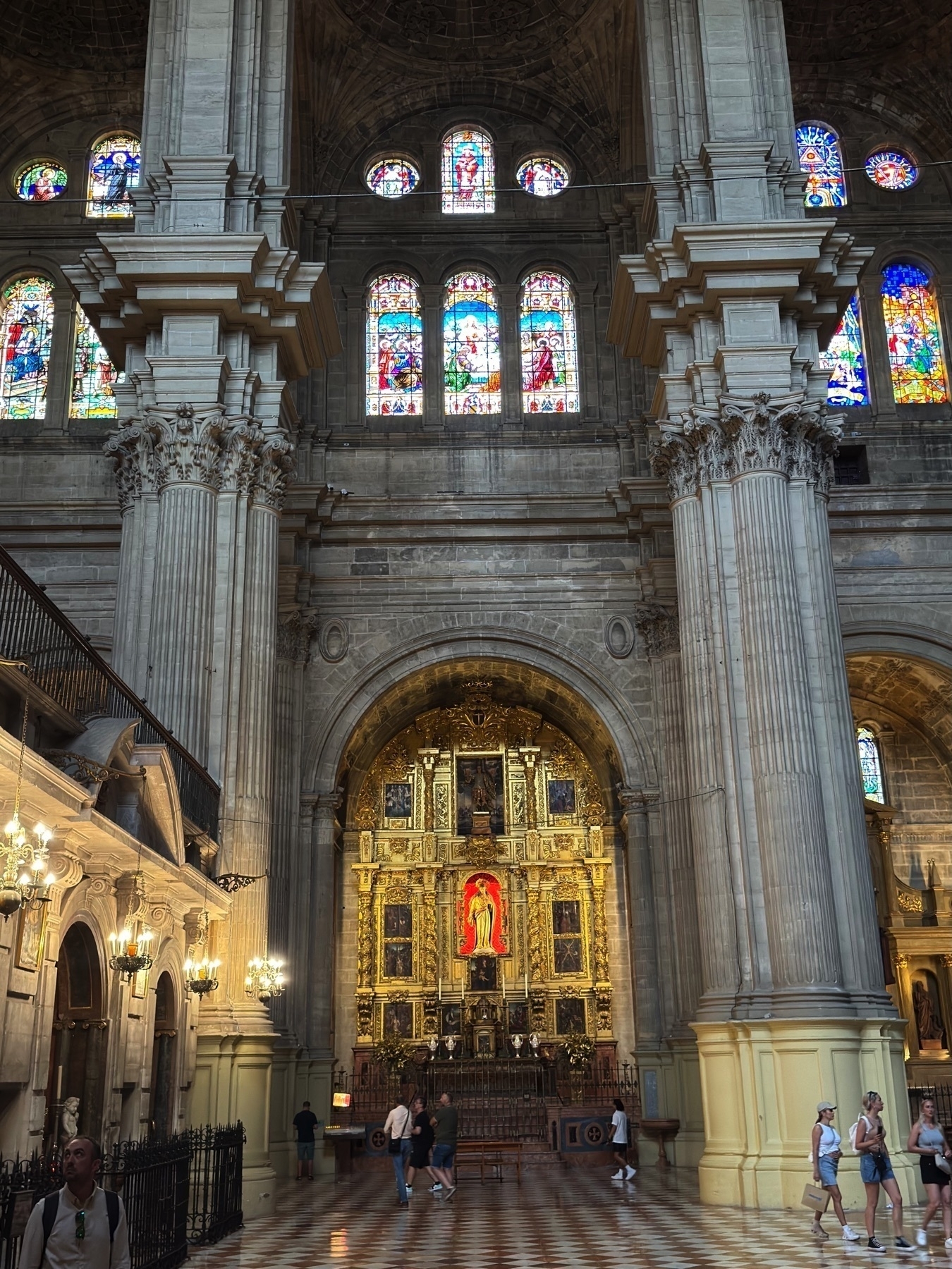 A golden altarpiece illuminated under soft light, with a central figure of the Virgin Mary in vivid red, set among detailed carvings and religious paintings. Above, colorful stained glass windows allow natural light to spill into the cathedral’s spacious nave.