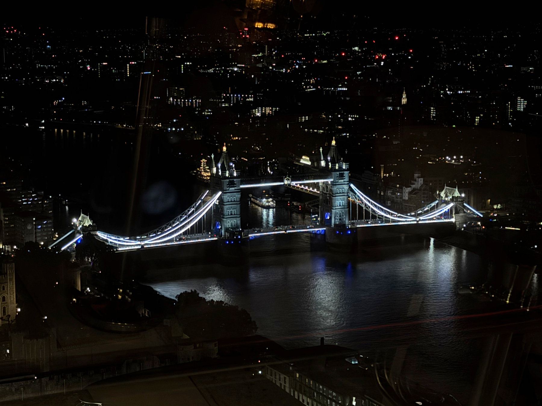 Tower Bridge lit in white and blue spans the Thames at night, its twin towers reflected in the dark water with surrounding city lights stretching into the distance.