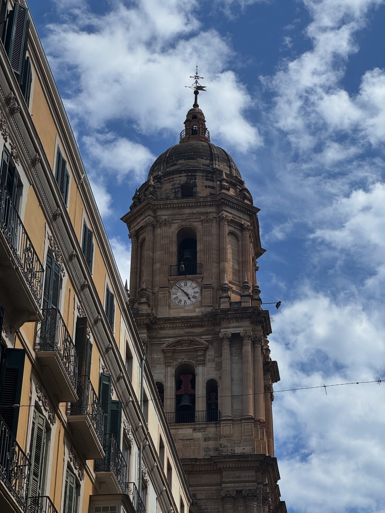View looking up at the Málaga Cathedral’s bell tower with a clock, framed on the left by traditional Spanish apartment buildings with wrought-iron balconies. The tall tower, with visible bells and ornate stone details, stands against a partly cloudy sky, capturing the architectural elegance and vertical prominence of the structure.