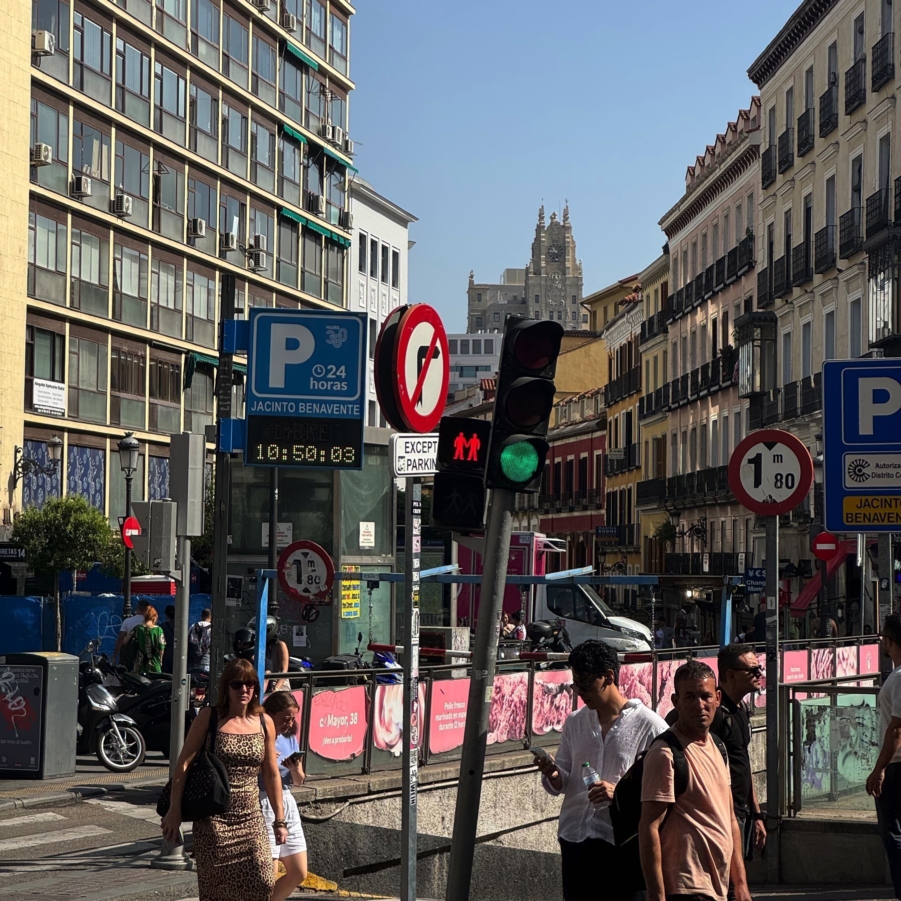 Urban intersection with a green pedestrian signal showing two red figures holding hands. The scene includes a mix of modern and traditional buildings, a digital parking sign, and a busy sidewalk with people crossing or walking in different directions.