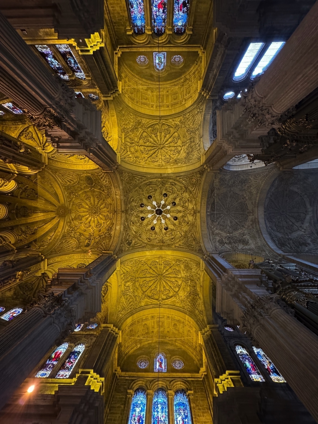 A dramatic upward view of the cathedral ceiling, showcasing elaborately carved golden vaults and ribbed domes, interspersed with stained glass windows. The intricate ceiling patterns glow under interior lighting, creating a breathtaking visual tapestry overhead.