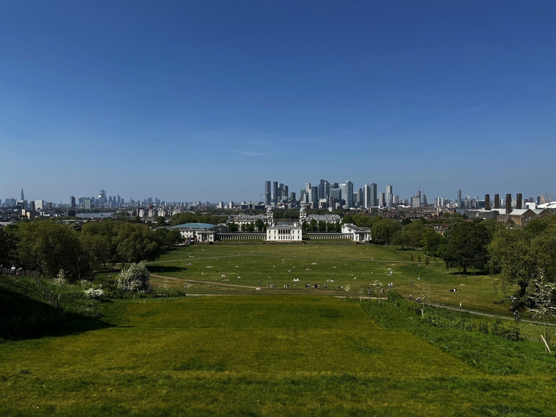 A panoramic view from Greenwich Park looking north over the Queen’s House and the Old Royal Naval College, with the Canary Wharf skyline rising in the distance. The wide green lawn below is dotted with people lounging and walking, and the city’s modern skyscrapers contrast with the classical architecture and open landscape in the foreground.