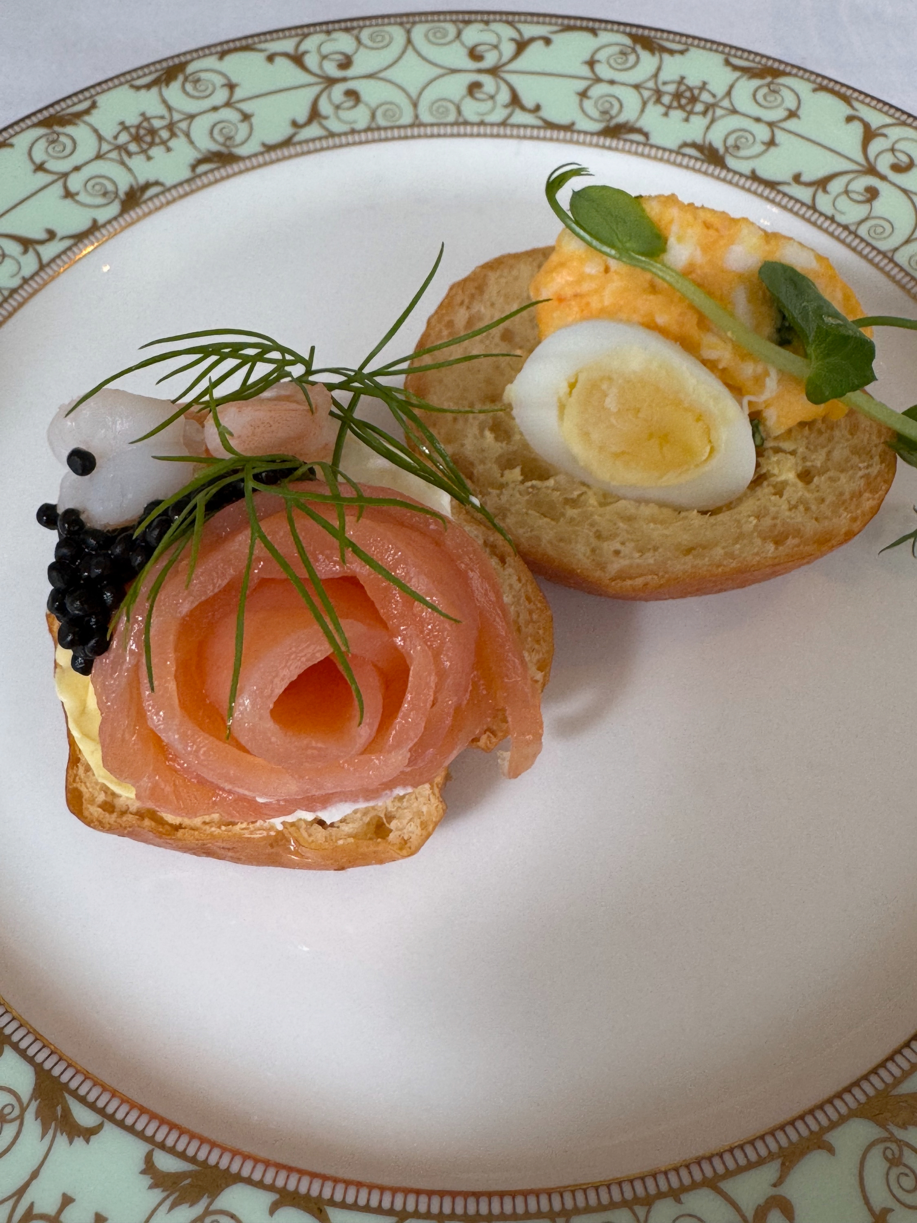 Two open-faced savory bites on a decorative plate: one topped with silky smoked salmon shaped like a rose, caviar, and fresh dill, the other with soft egg, creamy filling, and microgreens. The textures and colors look fresh, rich, and carefully balanced.