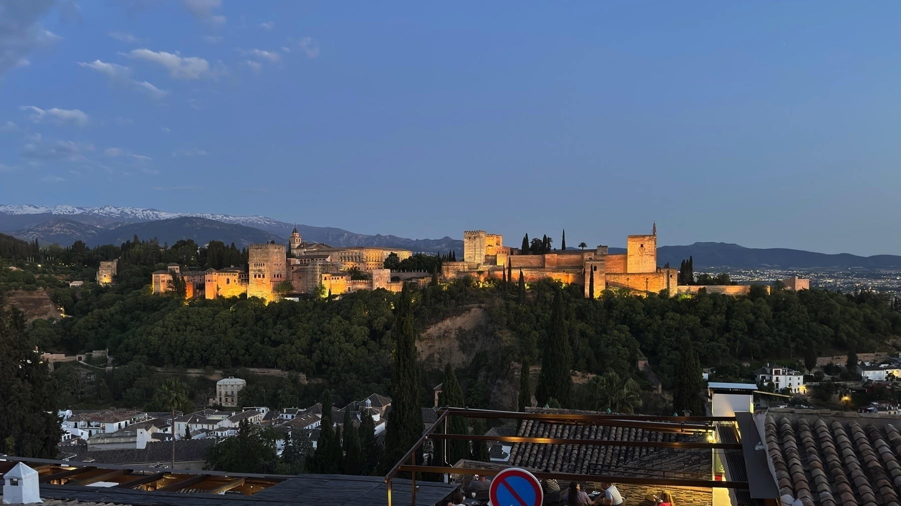 Evening view of the Alhambra fully illuminated, stretching across the hilltop with golden floodlights emphasizing its towers and walls. Snow-capped mountains are faintly visible in the background, and the rooftops of the Albaicín appear in the foreground below.