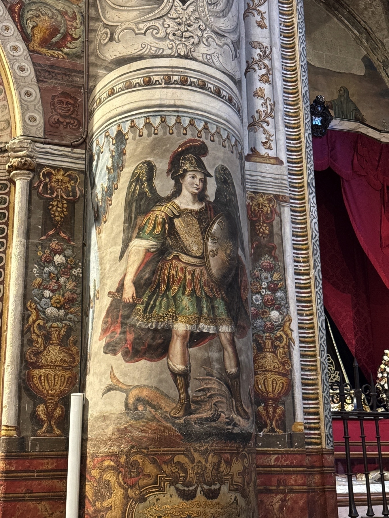 Fresco of a winged saint painted on a column, depicted in ornate armor holding a shield and sword while standing atop a defeated dragon. Surrounding floral motifs and decorative patterns enrich the scene, reflecting the artistic detail throughout the church interior.