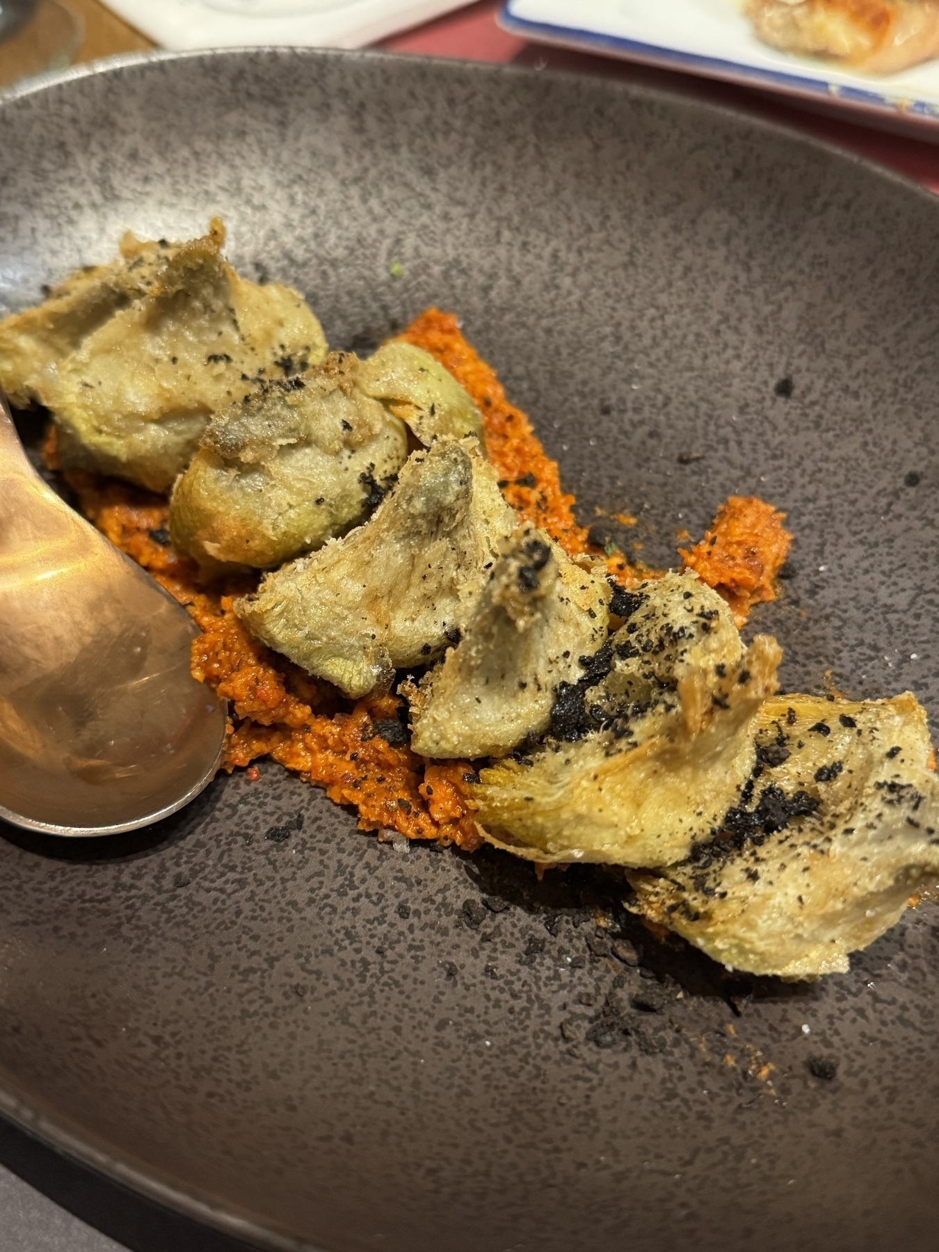 Fried artichoke hearts served over a bed of bright red romesco sauce and sprinkled with coarse black seasoning in a dark ceramic bowl.