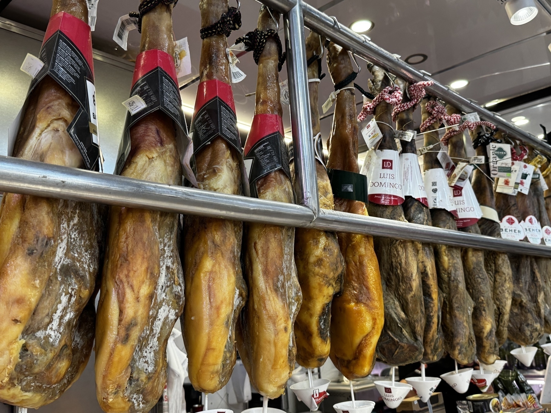 Close-up of a market stall inside Mercat Central de València displaying rows of hanging cured jamón legs with labeled tags and black or red rope ties, arranged on a metal rack.
