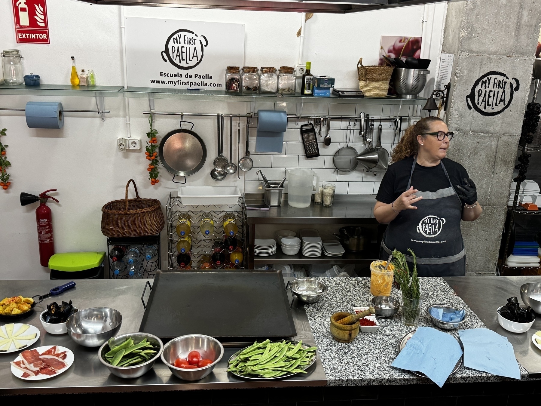 An instructor at My First Paella explaining the process, with paella ingredients like tomatoes, green beans, garlic, and meats arranged neatly in bowls on the counter.