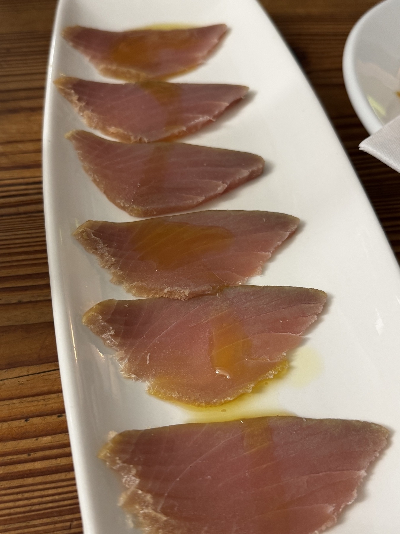 Six thin slices of smoked tuna laid out in a neat row on a long white plate, each piece lightly drizzled with golden oil.
