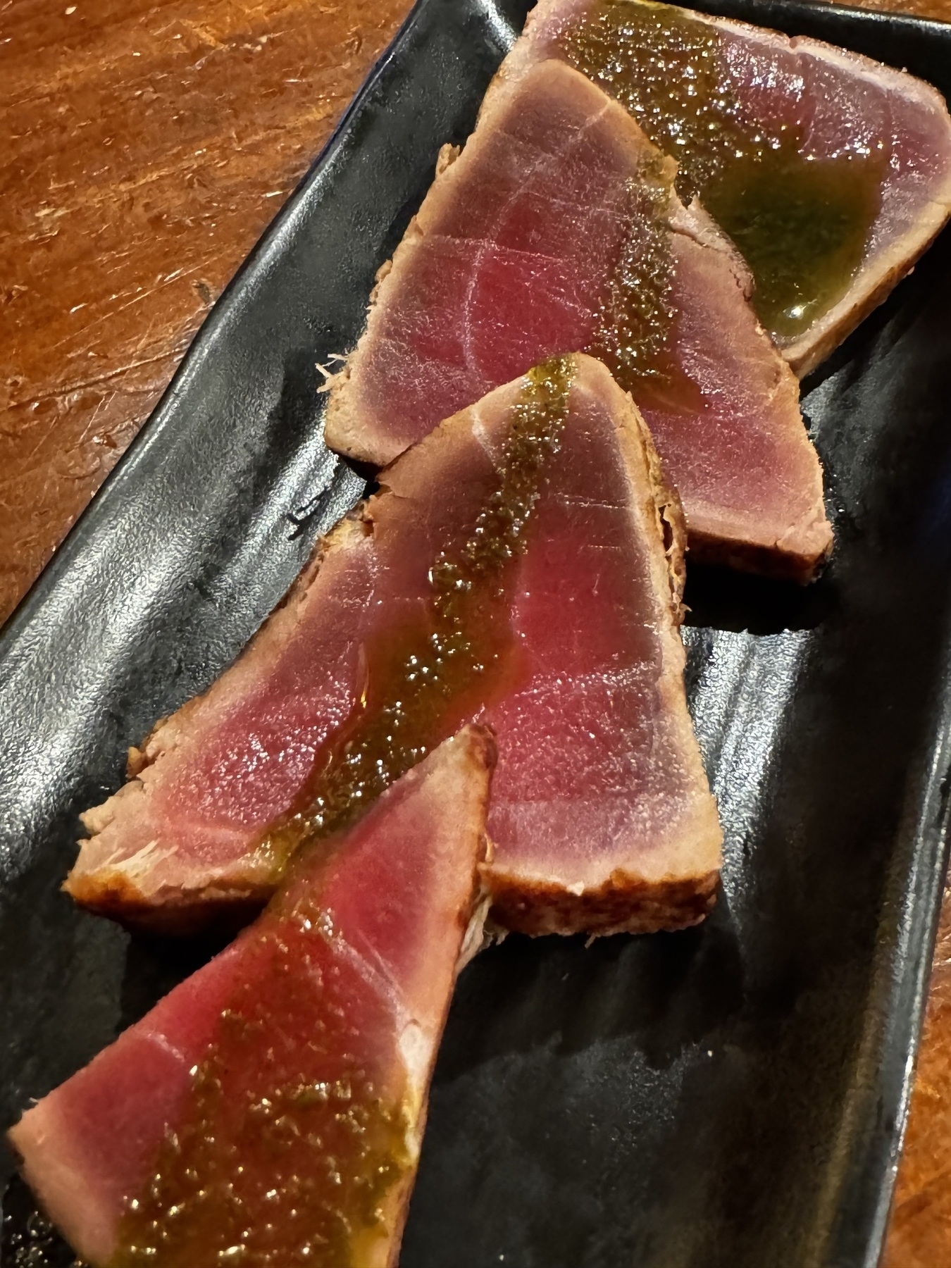 A black plate with thick slices of seared tuna, identified as Atún Corte Oriental. The tuna is raw and vivid pink at the center, with a lightly seared outer edge. A line of Asian-inspired sauce runs across the top, enhancing the rich, buttery look of the fish.