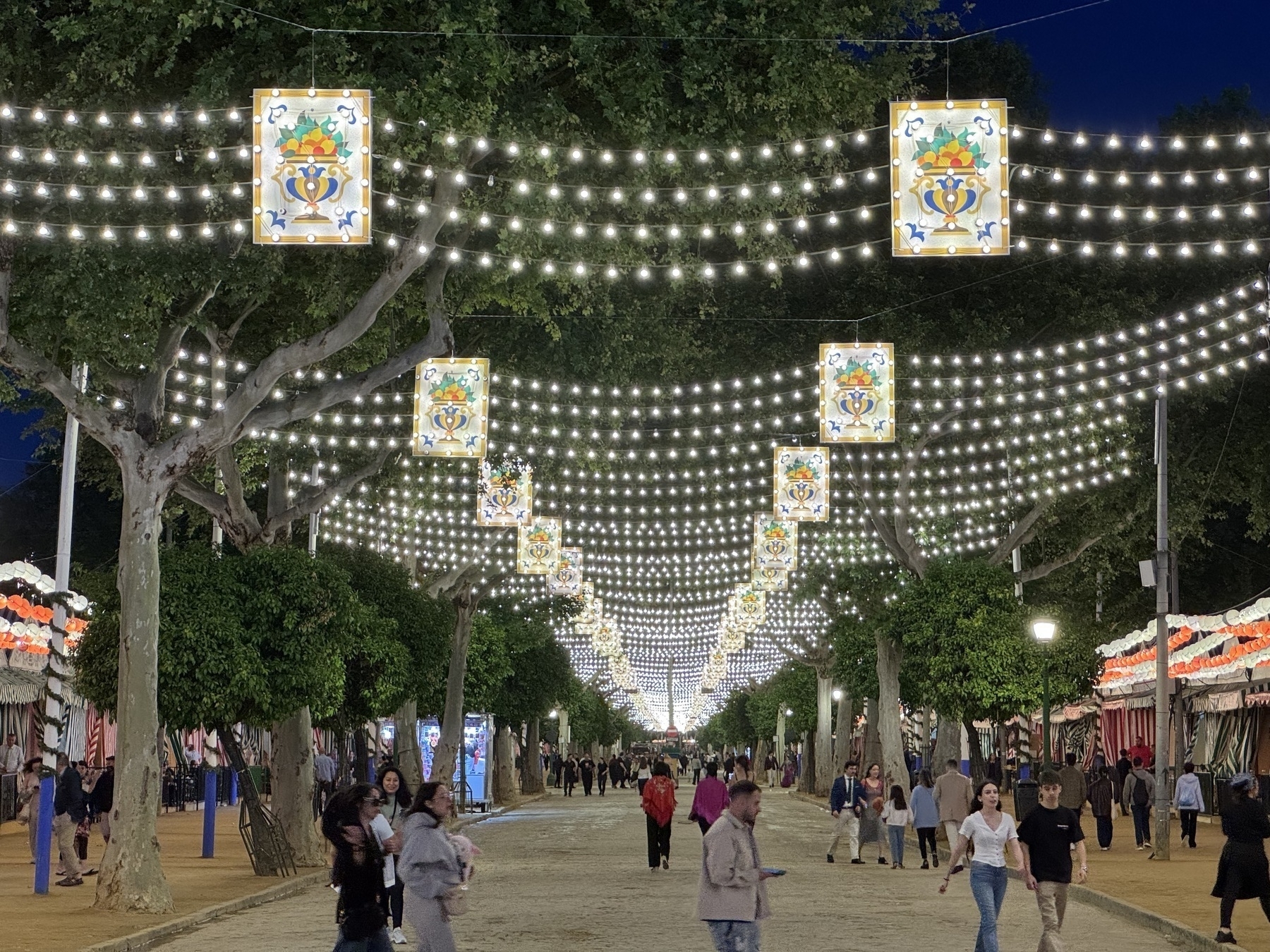 A wide pedestrian avenue is illuminated by rows of white string lights and decorative panels, suspended between trees lining the path. People stroll beneath the lights, creating a magical and orderly nighttime setting.