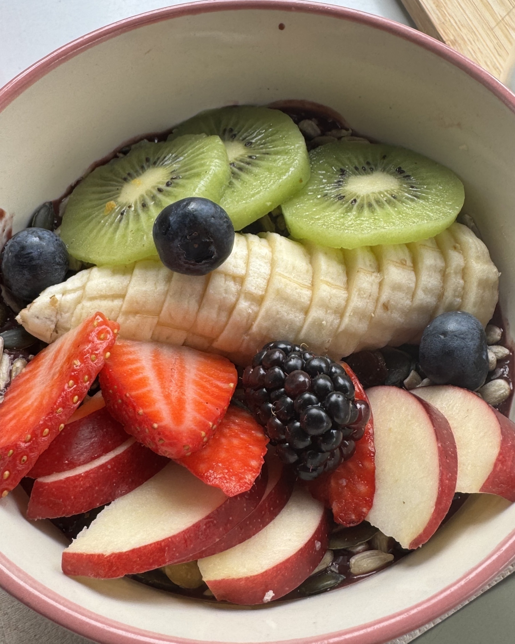 Overhead view of an açaí bowl from Feria 83, topped with artfully arranged fresh fruit. Slices of banana, kiwi, strawberries, and apple fan out over a dark purple açaí base, accented by whole blueberries and a glossy blackberry. The vibrant colors and textures—smooth banana, juicy berries, and crisp apple—make the bowl look both healthy and deliciously refreshing.