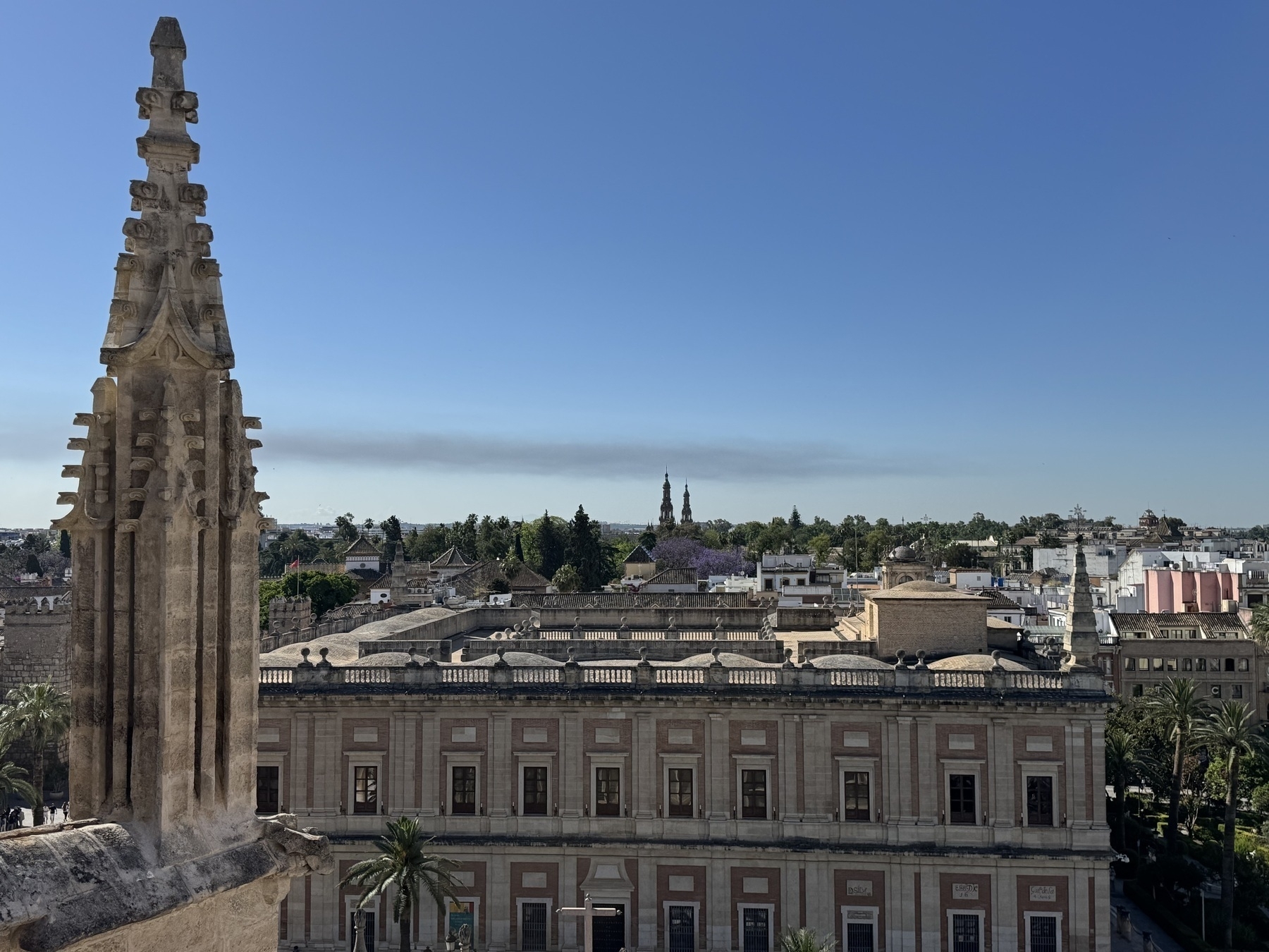 View from the cathedral’s rooftop showing a detailed Gothic pinnacle in the foreground and the rectangular, neoclassical Archivo General de Indias building below, with tree-lined cityscape and distant towers on the horizon.