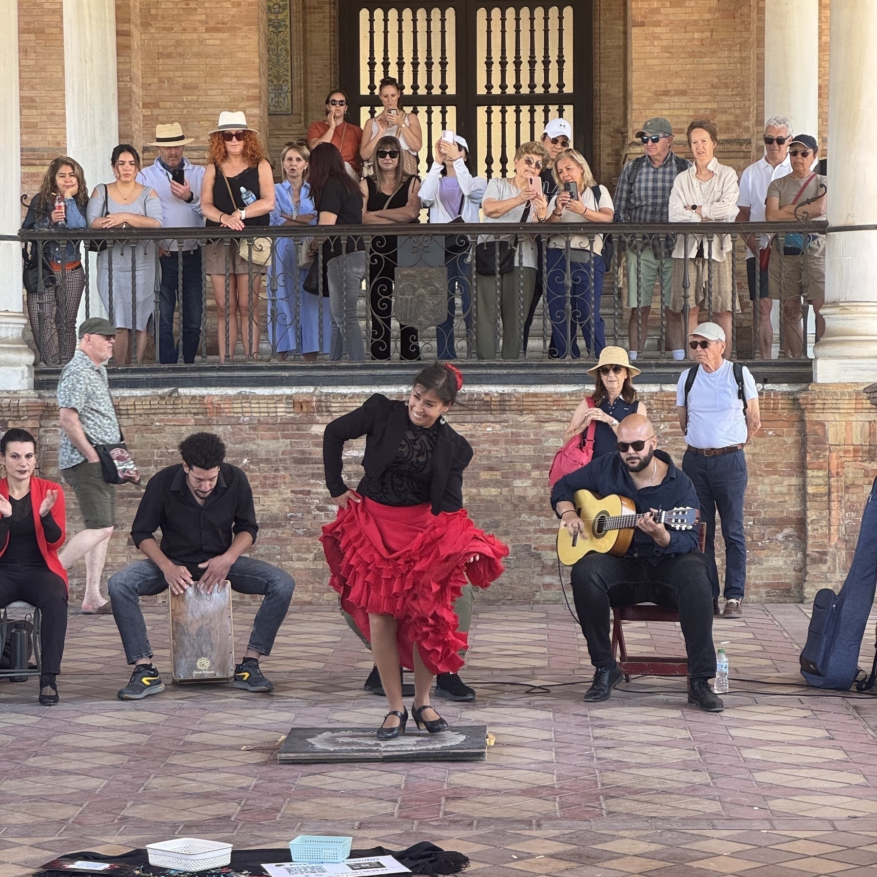 A passionate flamenco dancer in a red ruffled skirt and black top performs on a platform, mid-step, while a guitarist and percussionist accompany her. A diverse group of spectators watch from behind a railing under the arches of the building, many filming the show.