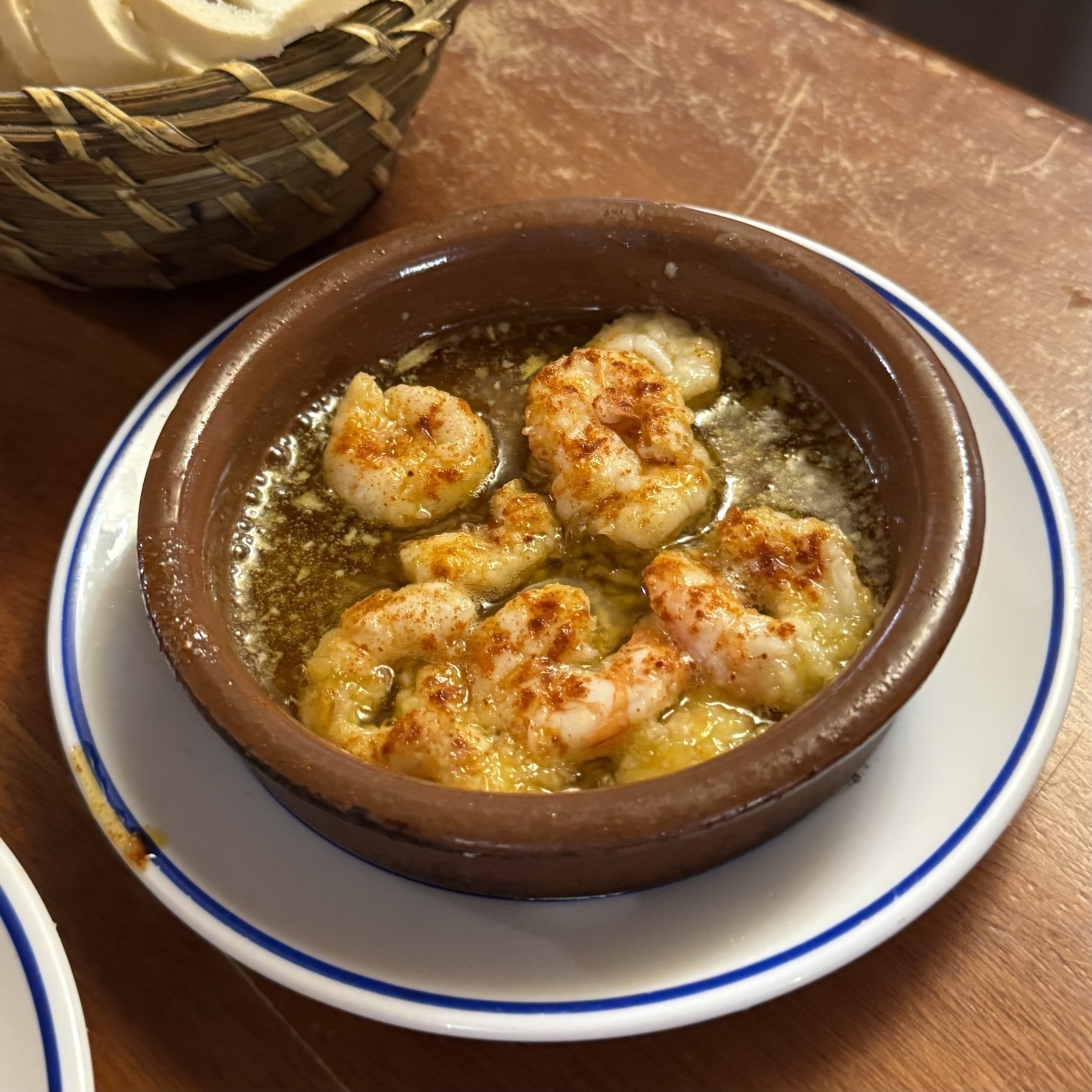 Earthenware dish filled with sizzling garlic shrimp in olive oil, topped with a sprinkling of paprika, with a basket of sliced bread nearby.