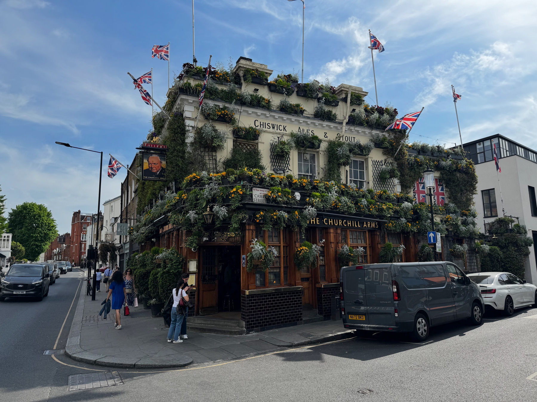 Corner pub covered in cascading greenery and yellow flowers, Union Jack flags flying from the roof and façade, with pedestrians and parked cars framing the street under a bright blue sky.