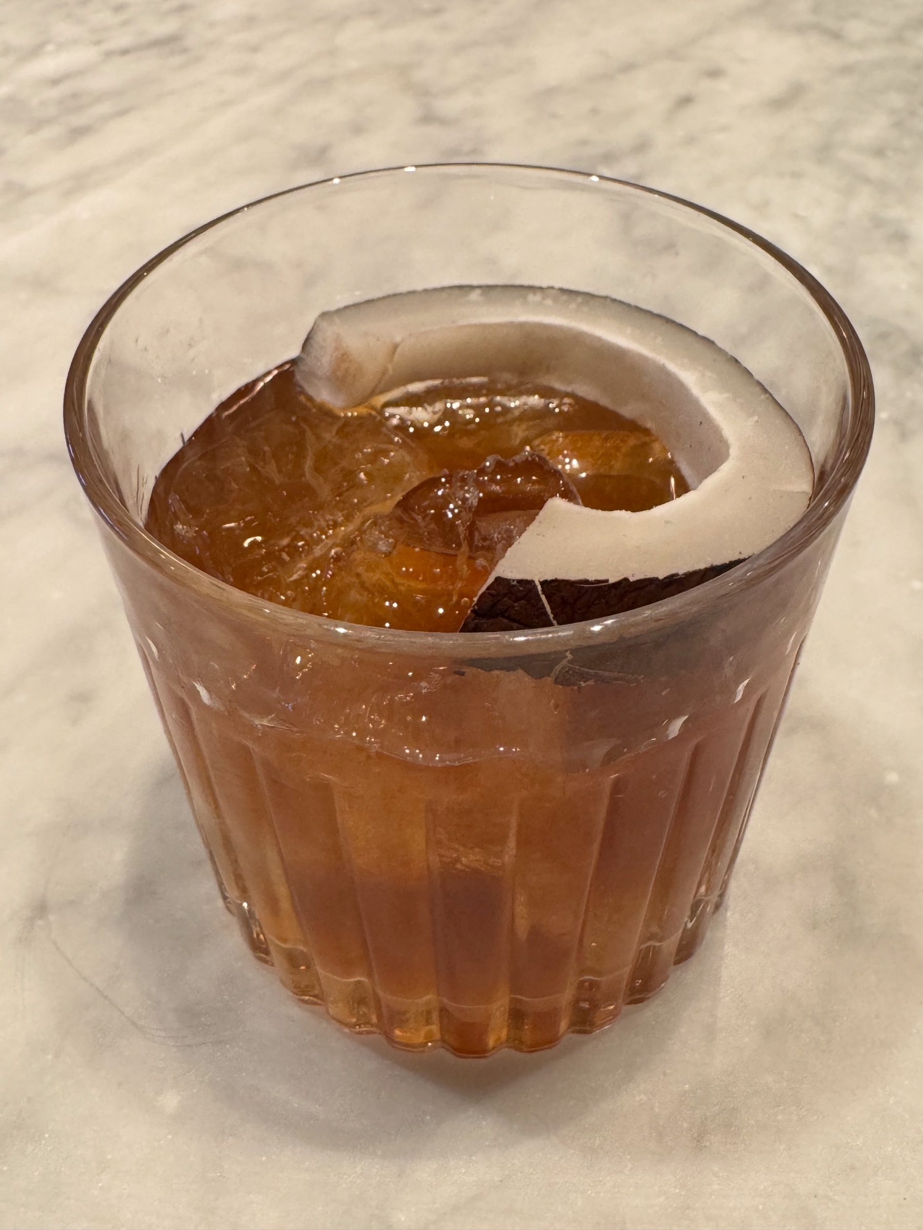 An amber-colored cocktail sits in a faceted rocks glass packed with ice, garnished with a wide curl of citrus peel. The drink catches the light, highlighting its clarity and depth of color against a pale marble surface, giving it a refined and refreshing appearance.