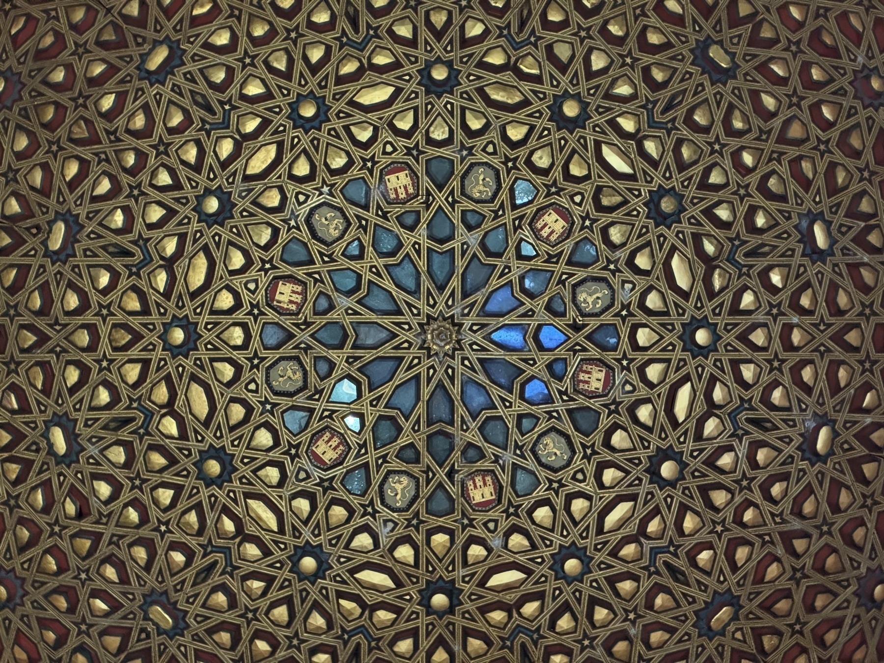 Close-up of an ornate coffered ceiling showcasing a symmetrical starburst design made of interlocking geometric shapes in gold, red, and blue. Small coats of arms and floral motifs are embedded in the pattern, highlighting the influence of Mudéjar art and craftsmanship.