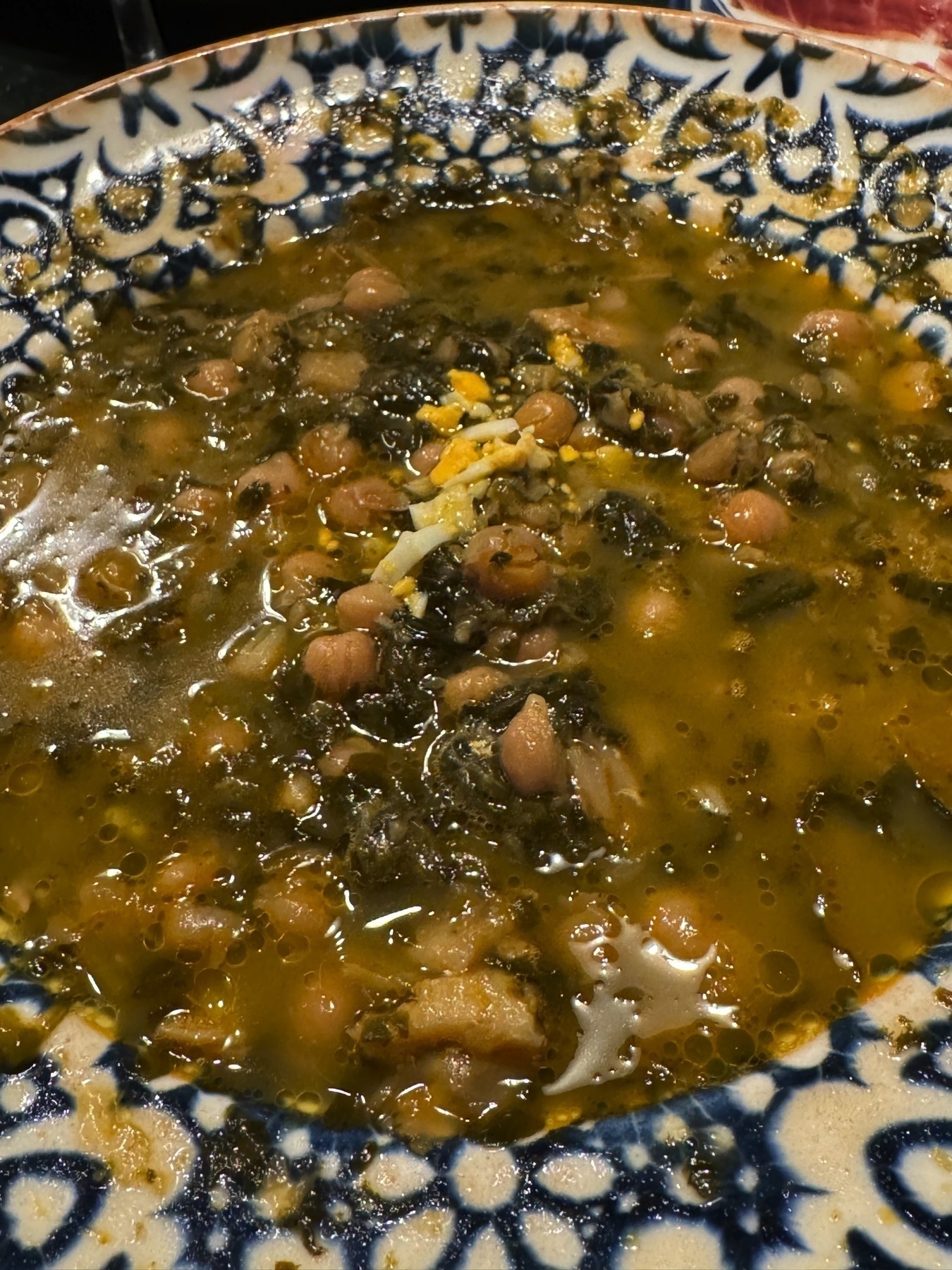A bowl of garbanzos con espinacas y bacalao, a traditional Cádiz dish made with chickpeas, spinach, and salted cod. The stew is rich and hearty, with visible chunks of flaked fish and greens in a glistening golden broth. A garnish of chopped boiled egg adds visual texture, and the patterned ceramic bowl enhances its rustic presentation.