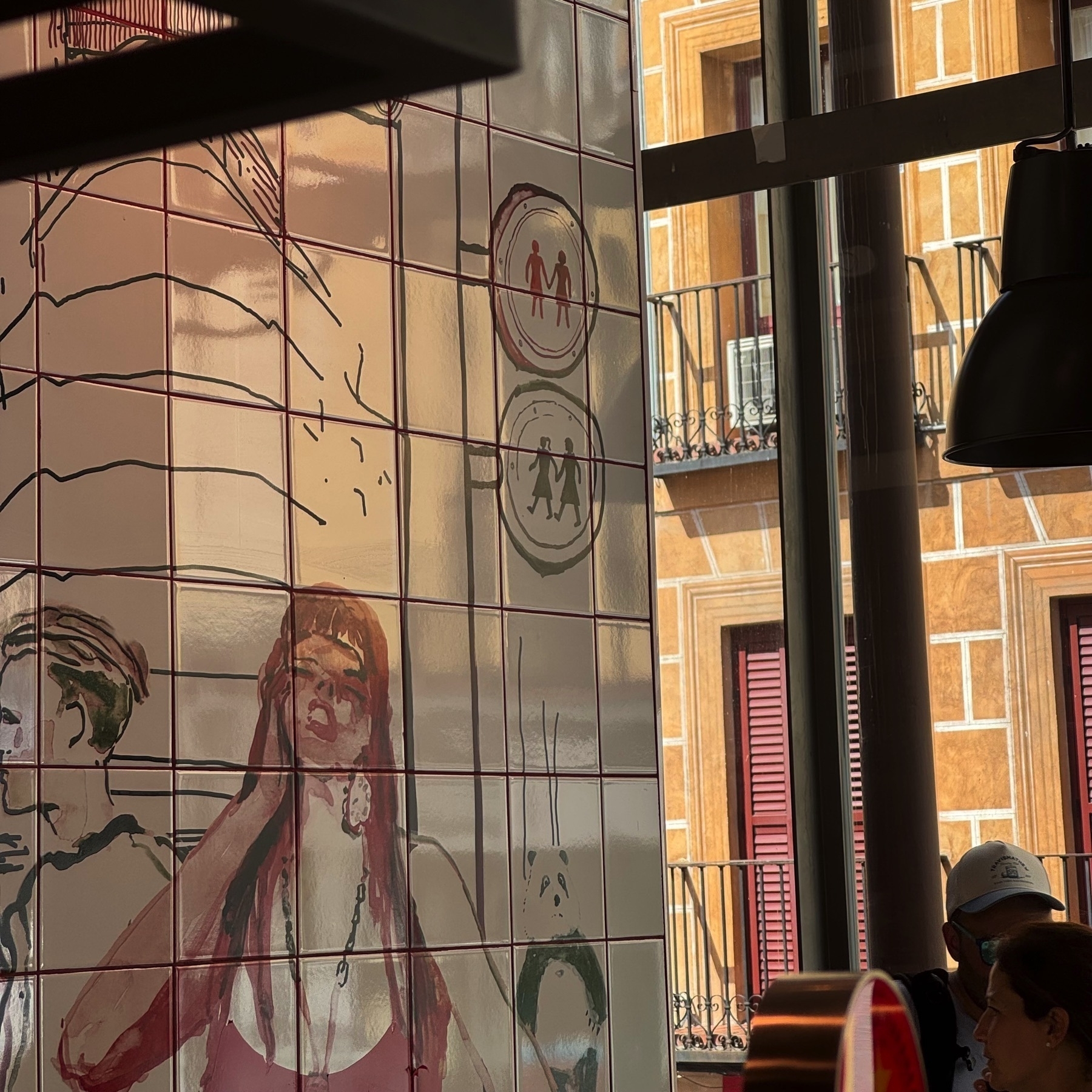A wall mural made of glossy ceramic tiles featuring a loosely sketched illustration of a red-haired woman in a tank top, holding her hand to her head. The scene includes minimalistic figures and stylized street signs, with reflections of the market’s interior and a view of the building across the street visible through the adjacent window.