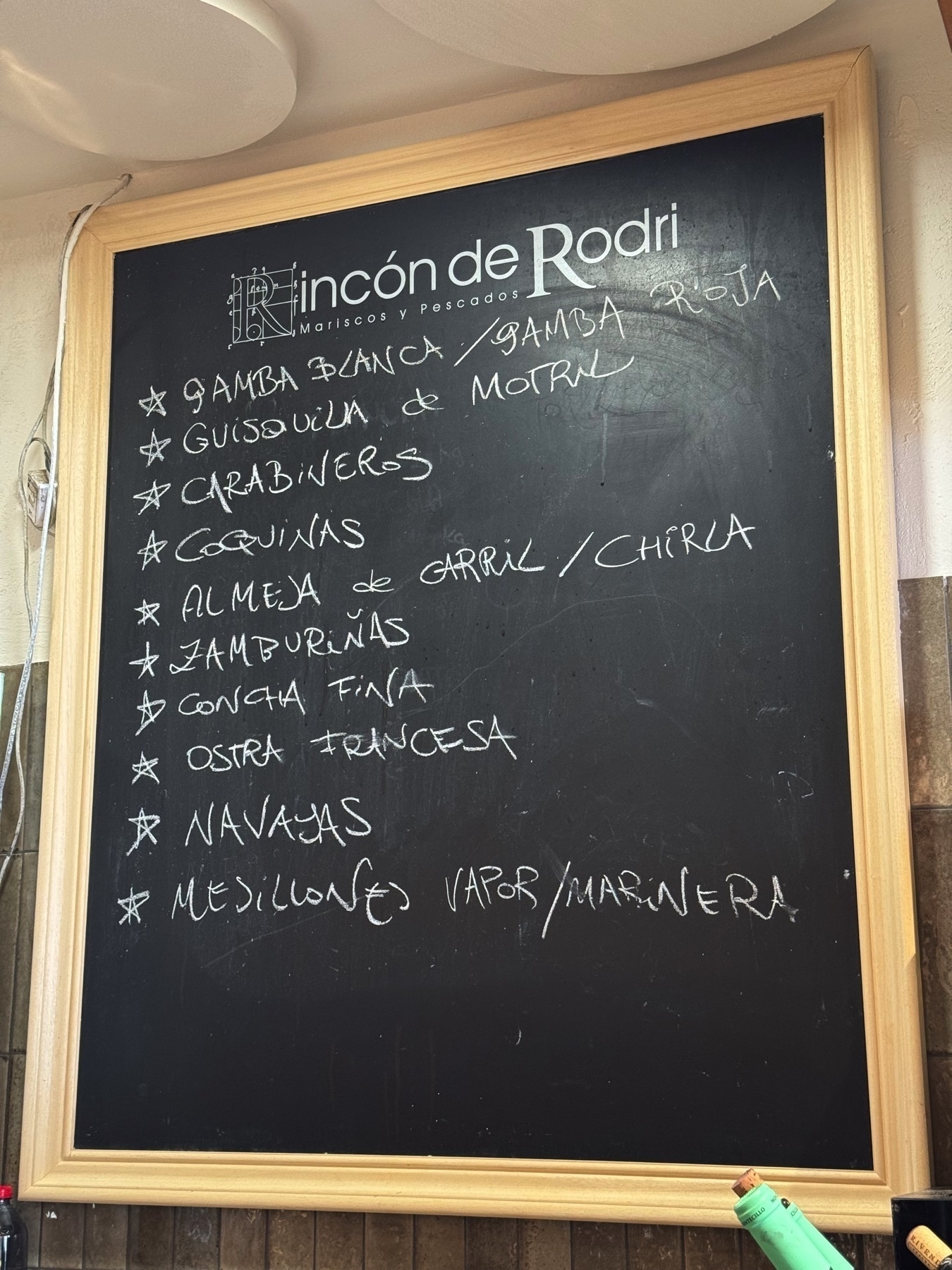 A chalkboard menu listing fresh seafood offerings in handwritten Spanish, framed in light wood. Items include gamba blanca, carabineros, zamburiñas, and ostras, with stars highlighting select dishes. The casual script and simple presentation evoke a sense of local charm and authenticity.