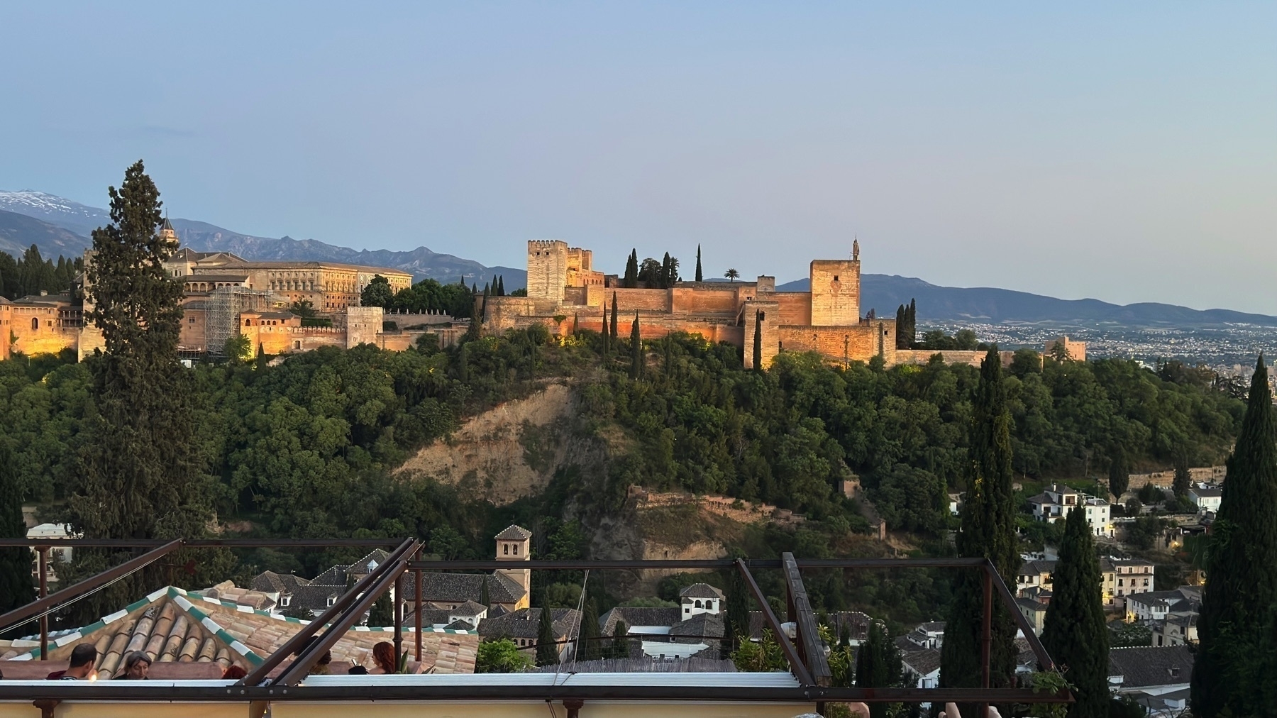 Panoramic view of the Alhambra at sunset, bathed in warm light and framed by surrounding trees. A rooftop terrace with patrons is visible in the foreground, and the city of Granada extends into the distance under a soft, pale sky.