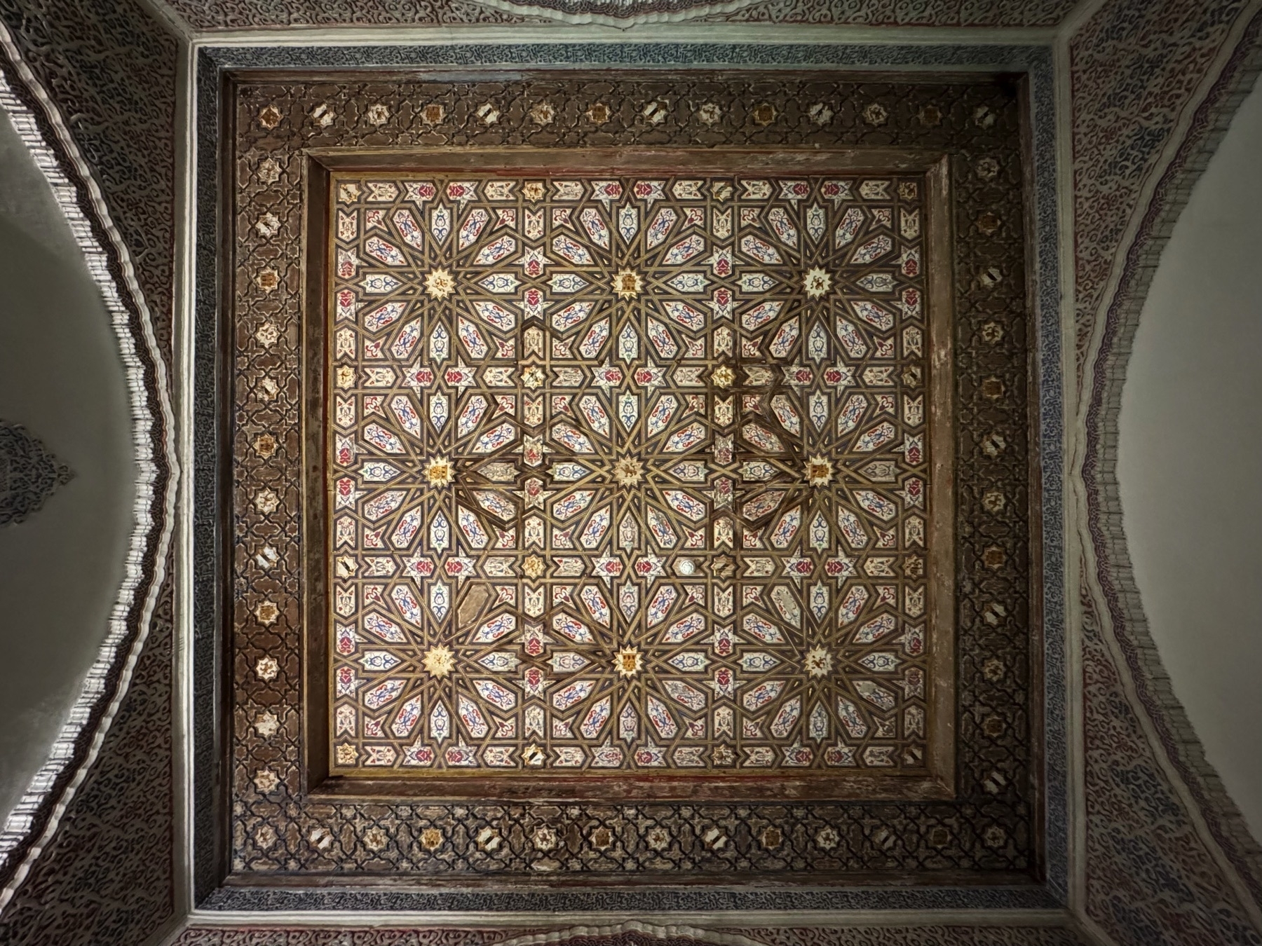 Artesonado ceiling with interlacing stars and floral patterns painted in red, white, and blue. The Mudéjar design uses wood and polychrome accents to create a symmetrical, geometric composition framed by carved borders.