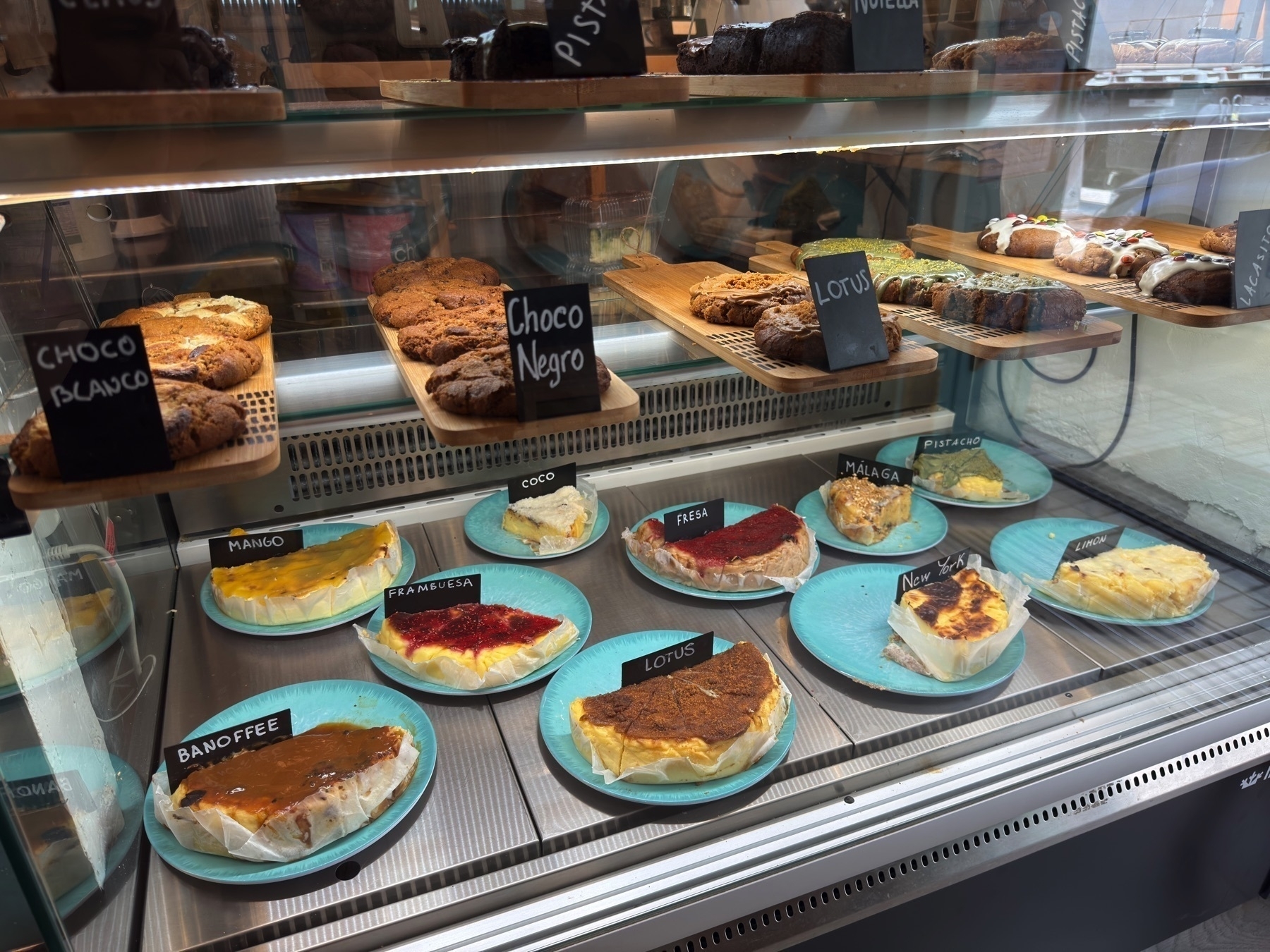 A dessert display filled with cheesecakes and baked goods labeled by flavor—like mango, fresa, lotus, and banoffee. The slices rest on turquoise plates in a chilled case, with cookies and other treats above them on wooden trays.