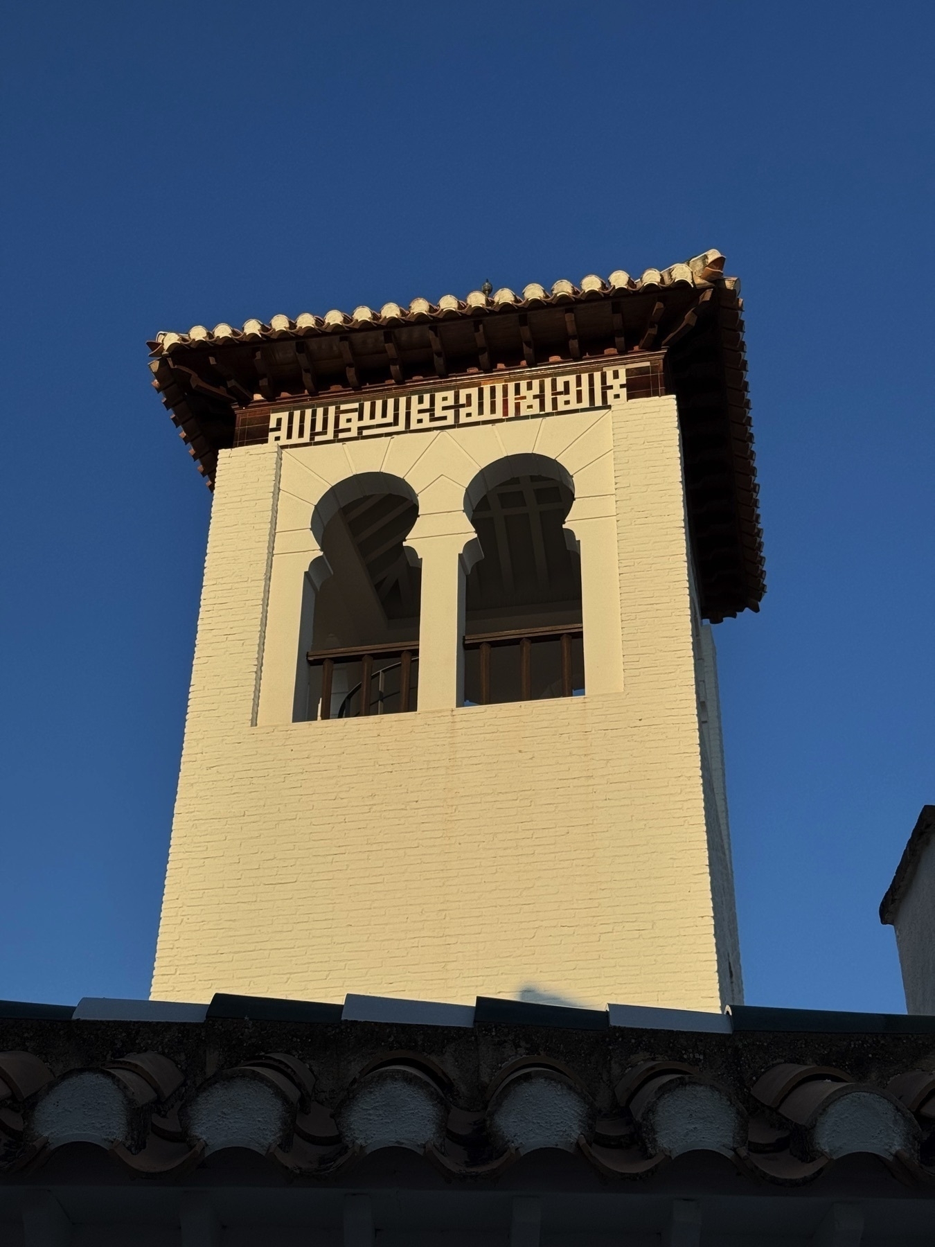 Close-up of a white brick bell tower with a pair of arched openings near the top and Arabic calligraphy-style ornamentation just below the roof. The warm evening light accentuates the texture of the walls and wooden roof beams.
