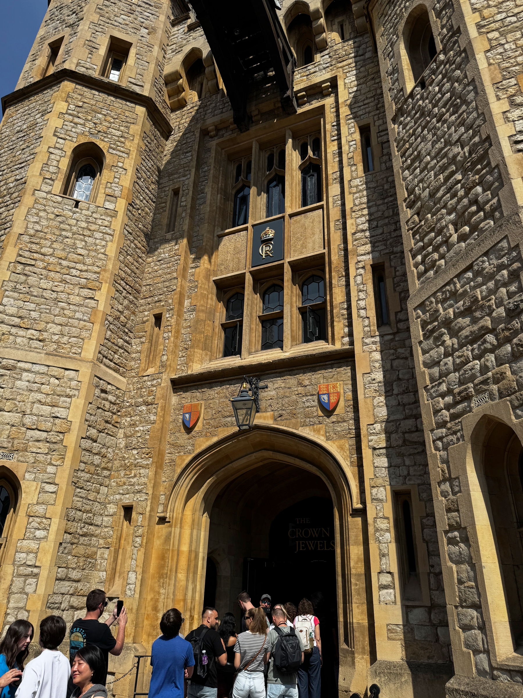 Visitors queue beneath a tall arched stone entrance marked by heraldic shields and a royal monogram, with sunlight highlighting the warm tones and textures of the medieval masonry around the doorway.