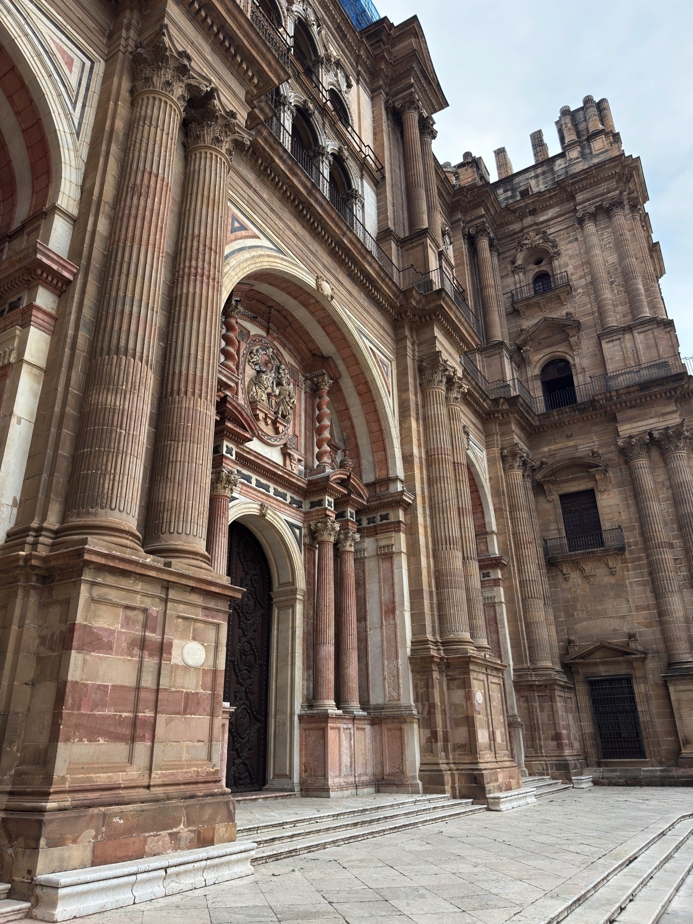 Close-up of the cathedral’s exterior facade, highlighting tall fluted columns, arched entrances, detailed stone carvings, and rose-colored marble accents. The classical architecture is imposing yet elegant against the stone plaza.