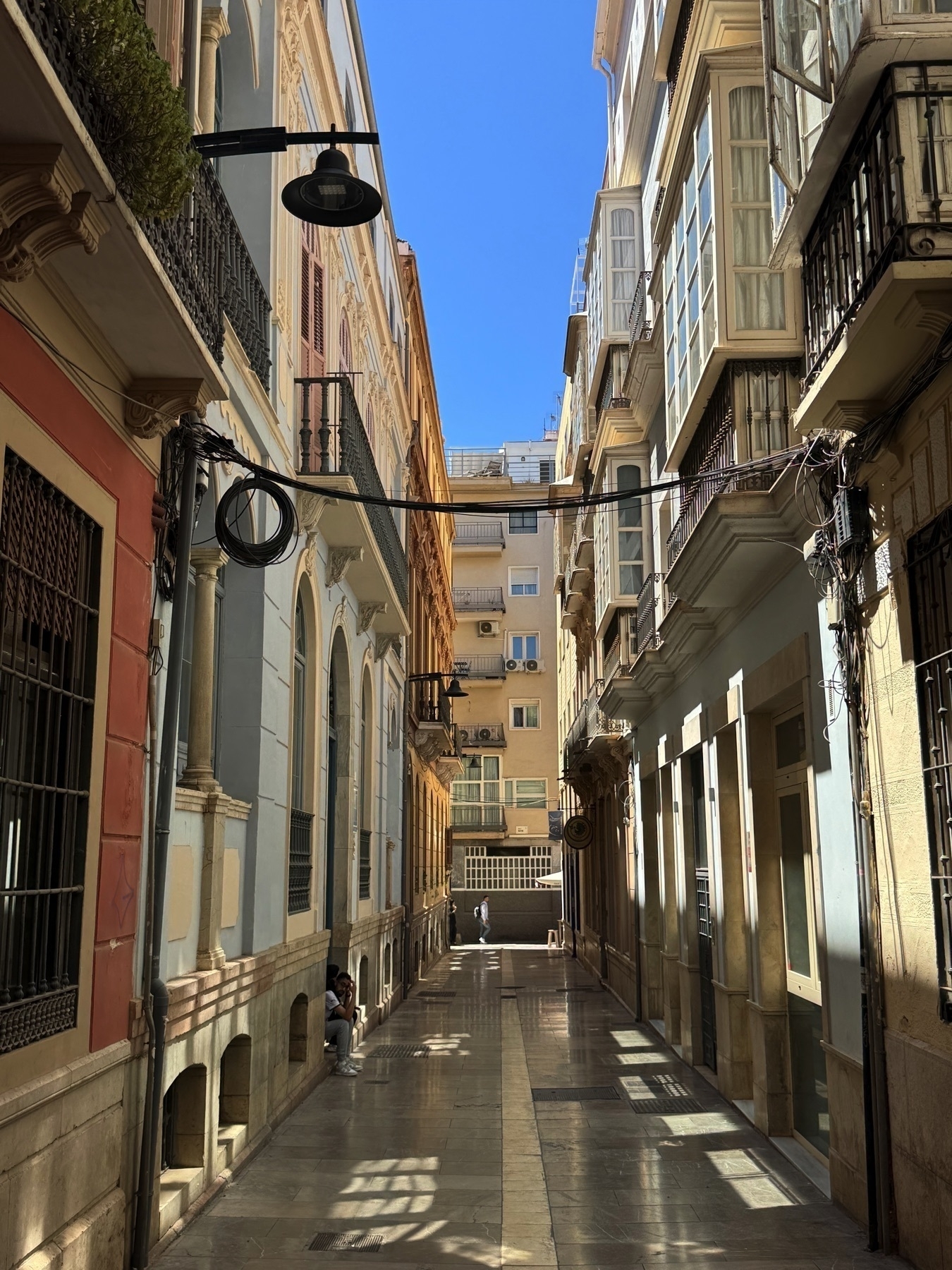 Quiet alleyway in Málaga’s old town lined with elegant 19th-century buildings in pastel tones. Sunlight reflects off polished stone pavement, and a person sits in a recessed doorway, adding a sense of calm and solitude.