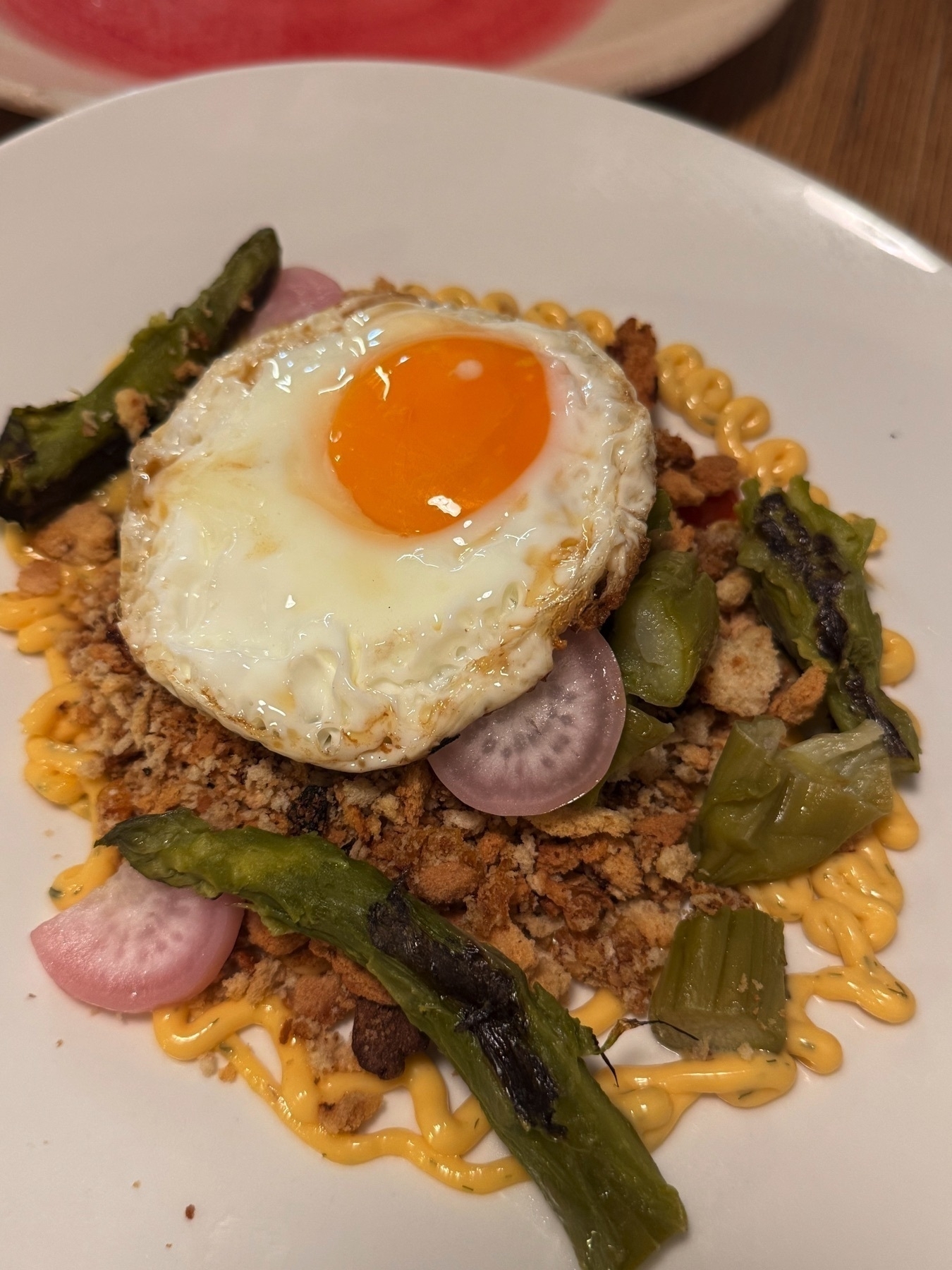 Artful plating of a sunny-side-up egg atop a bed of savory crumble, roasted peppers, pickled onions, and a zigzag of creamy sauce, showcasing a colorful and textural contrast.