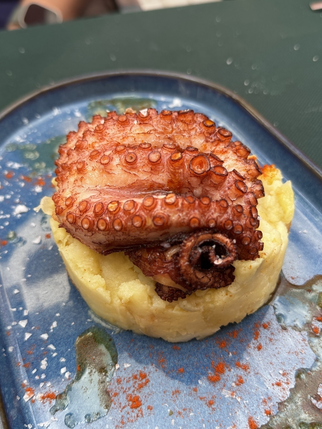 A dish of grilled octopus atop a bed of mashed potatoes, served on a blue ceramic plate with sea salt and paprika sprinkled around.