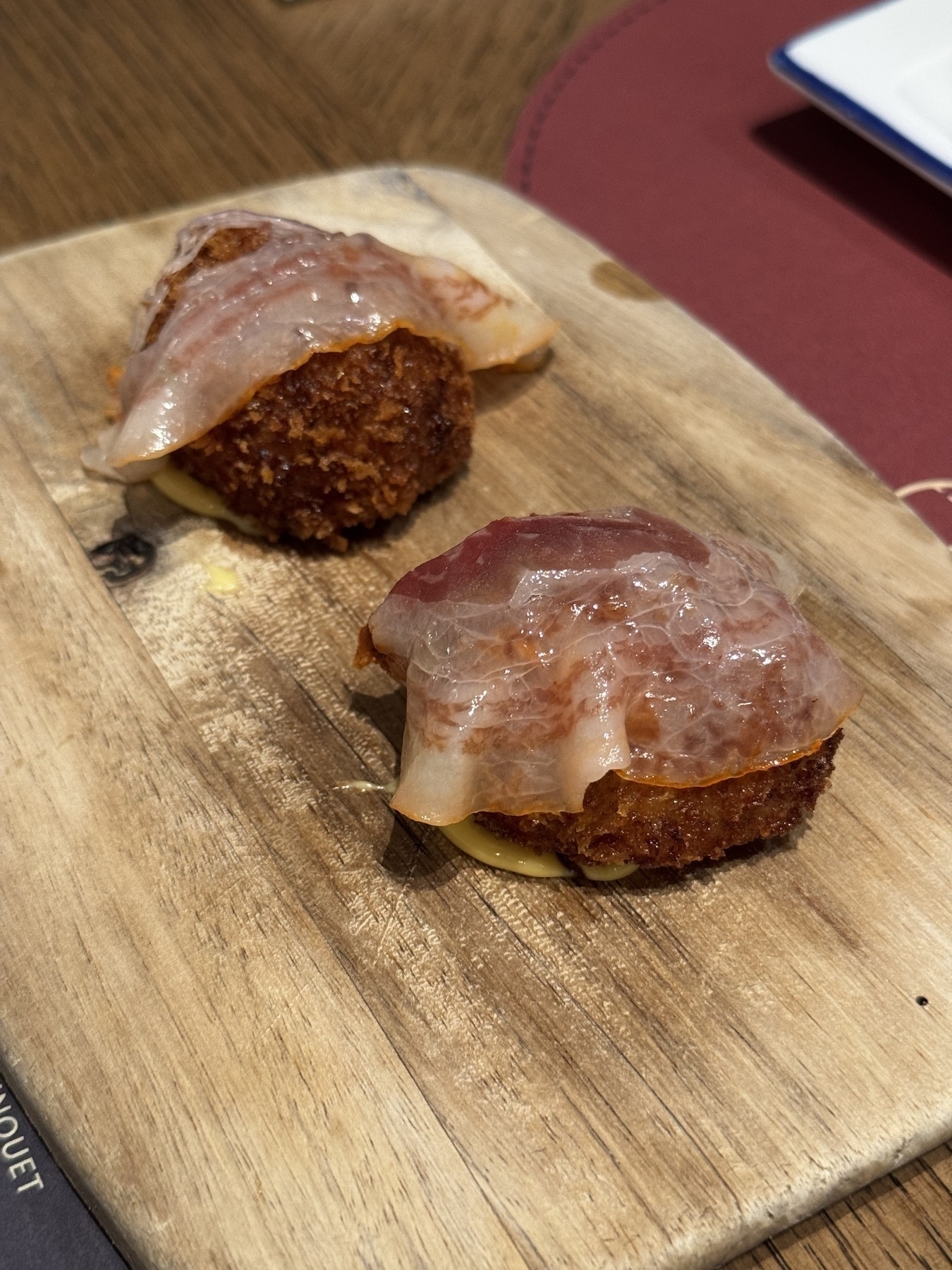 Two golden-brown croquettes topped with translucent slices of cured ham, plated on a wooden board with a touch of sauce underneath.