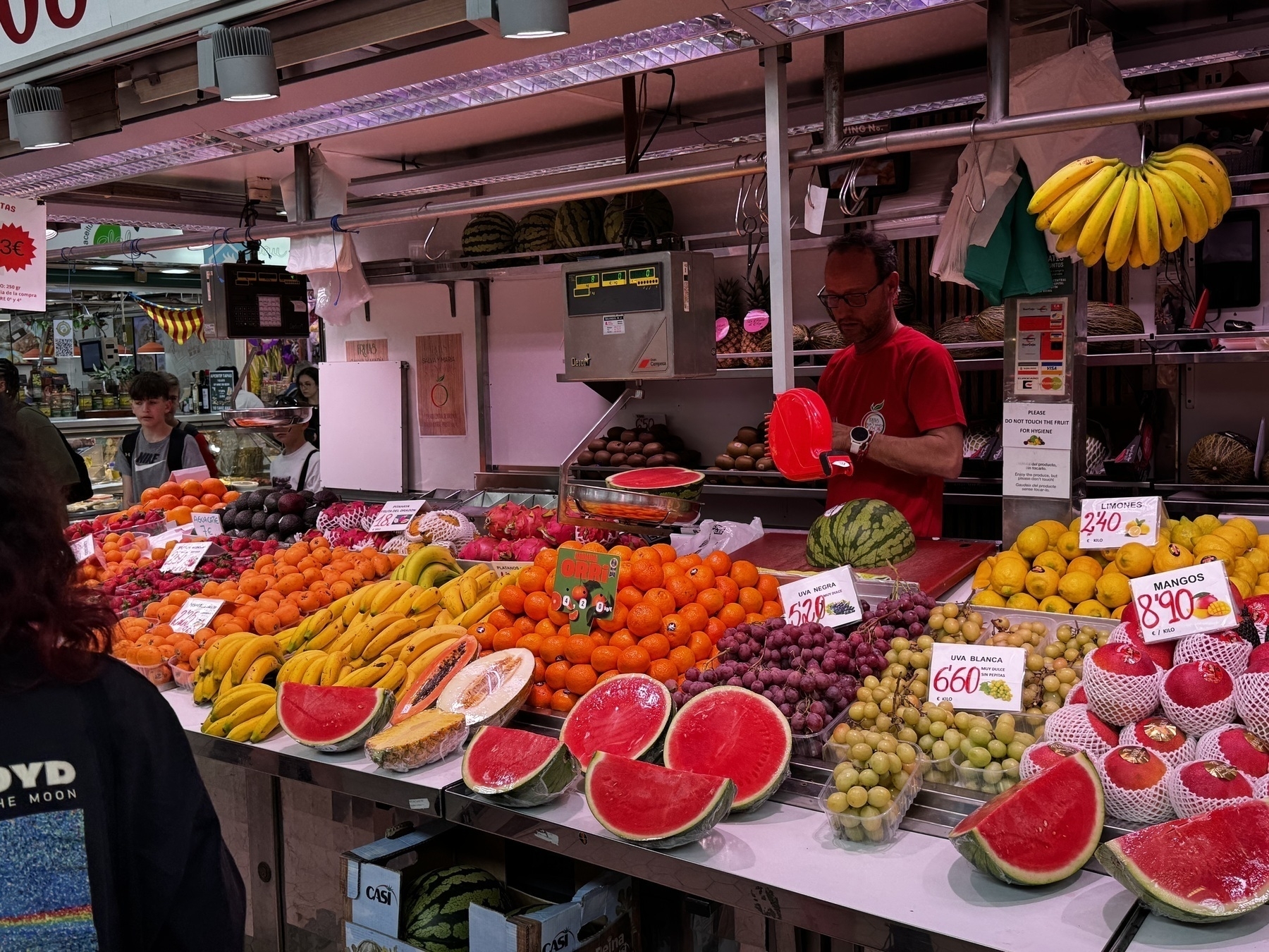 Fruit stand at Mercat Central de València with a vendor slicing watermelon behind a colorful display of fresh produce, including bananas, grapes, melons, citrus fruits, and mangoes, under bright market lighting.