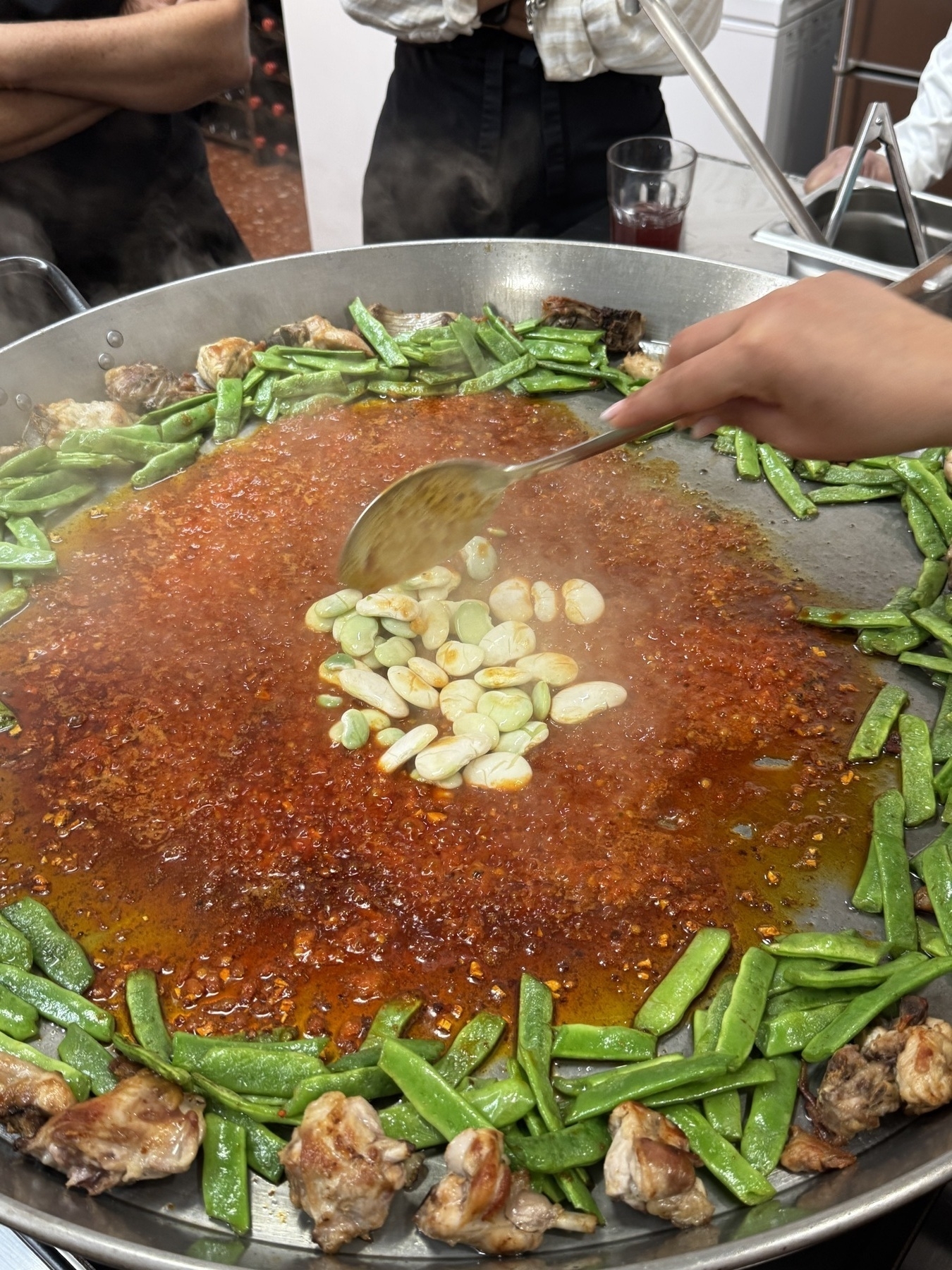 A hand ladling large white beans into a wide pan of simmering red broth, surrounded by chicken and green beans, during an early stage of making paella Valenciana.