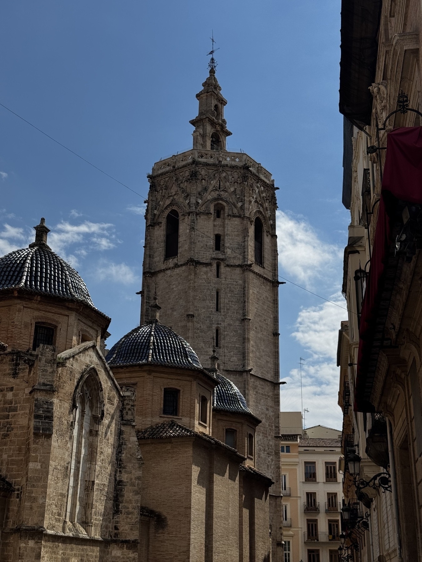 The Miguelete bell tower rises behind the tiled domes of a cathedral, its Gothic stonework dramatic against the sunlit sky. The structure’s height and weathered texture evoke centuries of architectural heritage.
