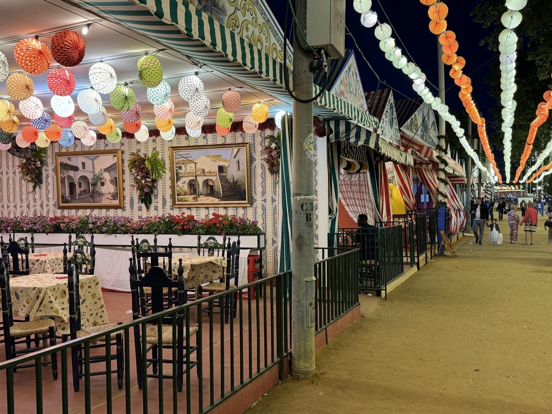 A row of colorful striped tents with traditional decor and hanging paper lanterns in red, orange, green, and yellow. One tent is open, revealing simple wooden tables and chairs, evoking a sense of intimate gathering and tradition.