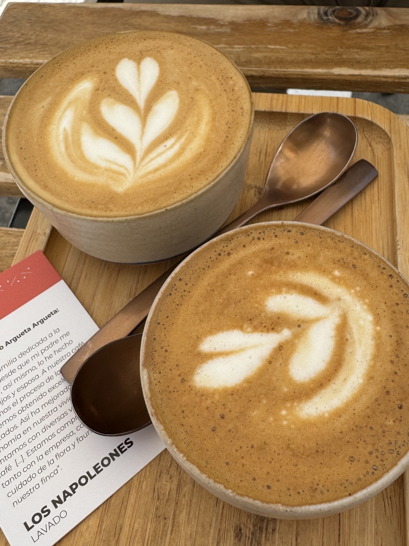 Two ceramic cups of latte on a wooden tray at Parcería Café, each topped with creamy, carefully poured latte art in leaf and tulip designs. The silky foam contrasts with the golden-brown espresso, and the warm tones of the tray and bronze spoons create an inviting atmosphere that emphasizes the care and quality in each cup.