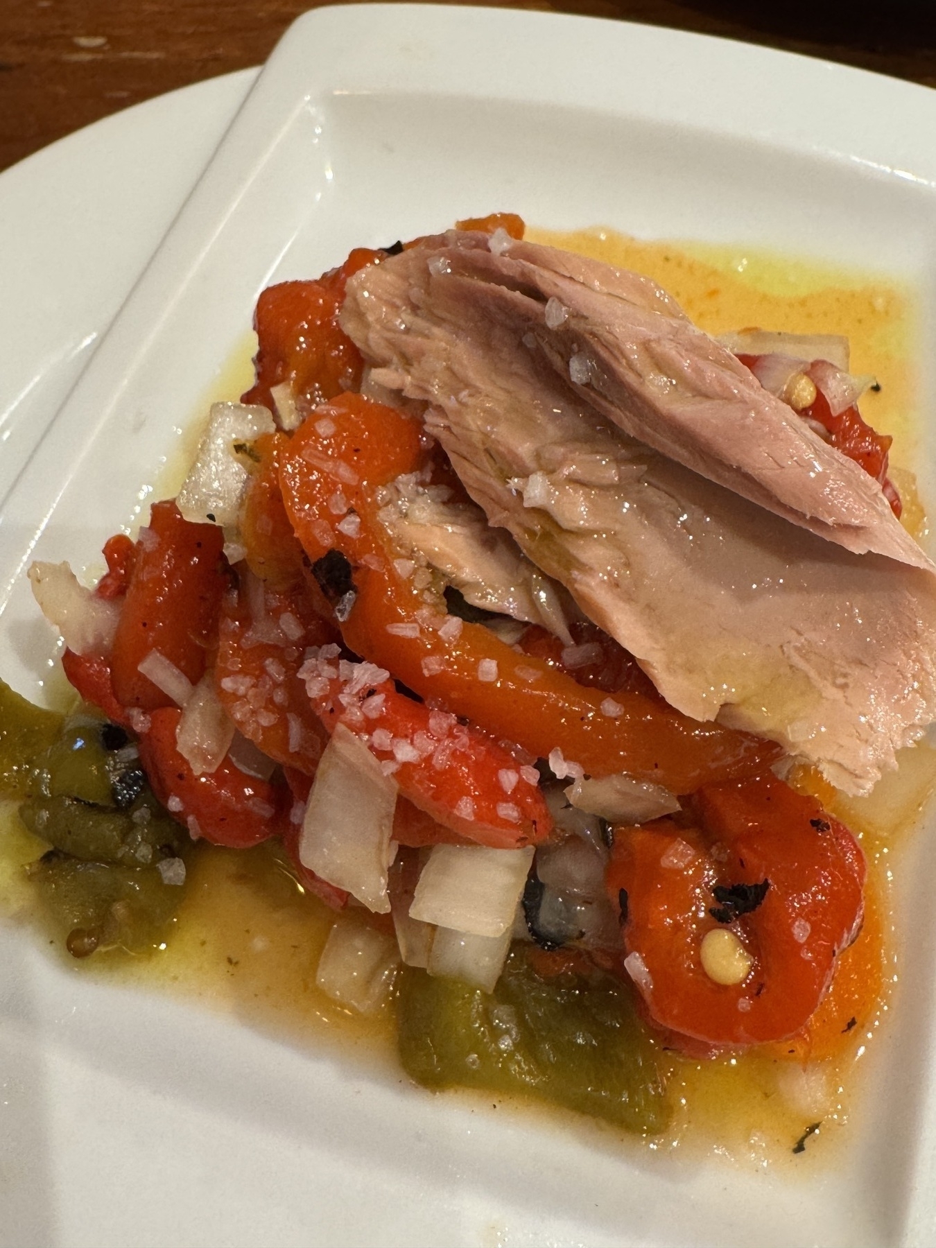 A white plate with Pimientos de la Axarquía—wood-fired roasted red peppers. The peppers are sliced and glistening with olive oil, mixed with diced white onions and sprinkled with coarse sea salt. Flakes of tuna or bonito rest on top, contrasting with the peppers’ intense color and shine.