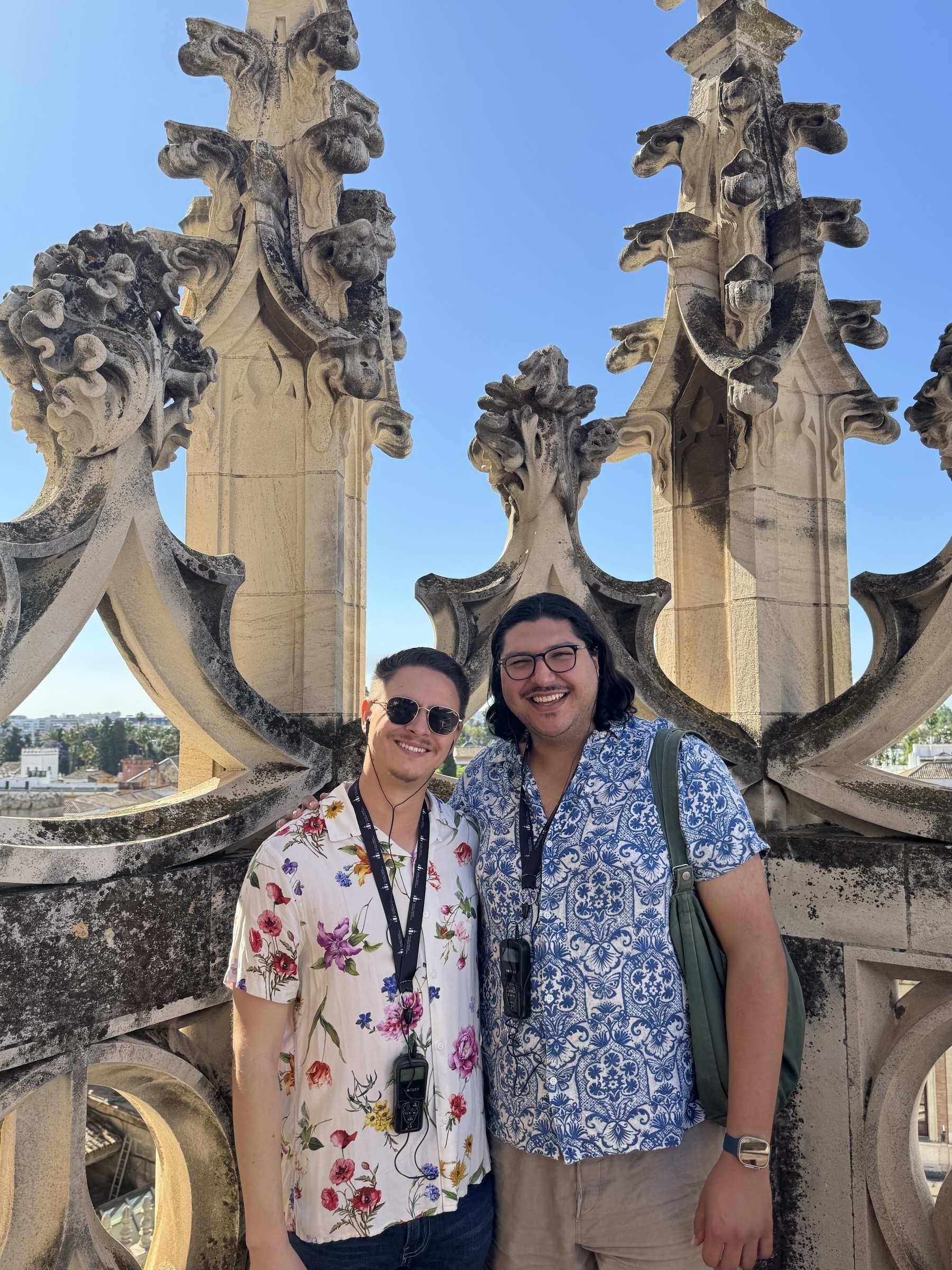Two smiling visitors standing close together in front of ornate stone balustrades with sculpted Gothic motifs, enjoying the rooftop tour with a clear blue sky and panoramic city views in the background.