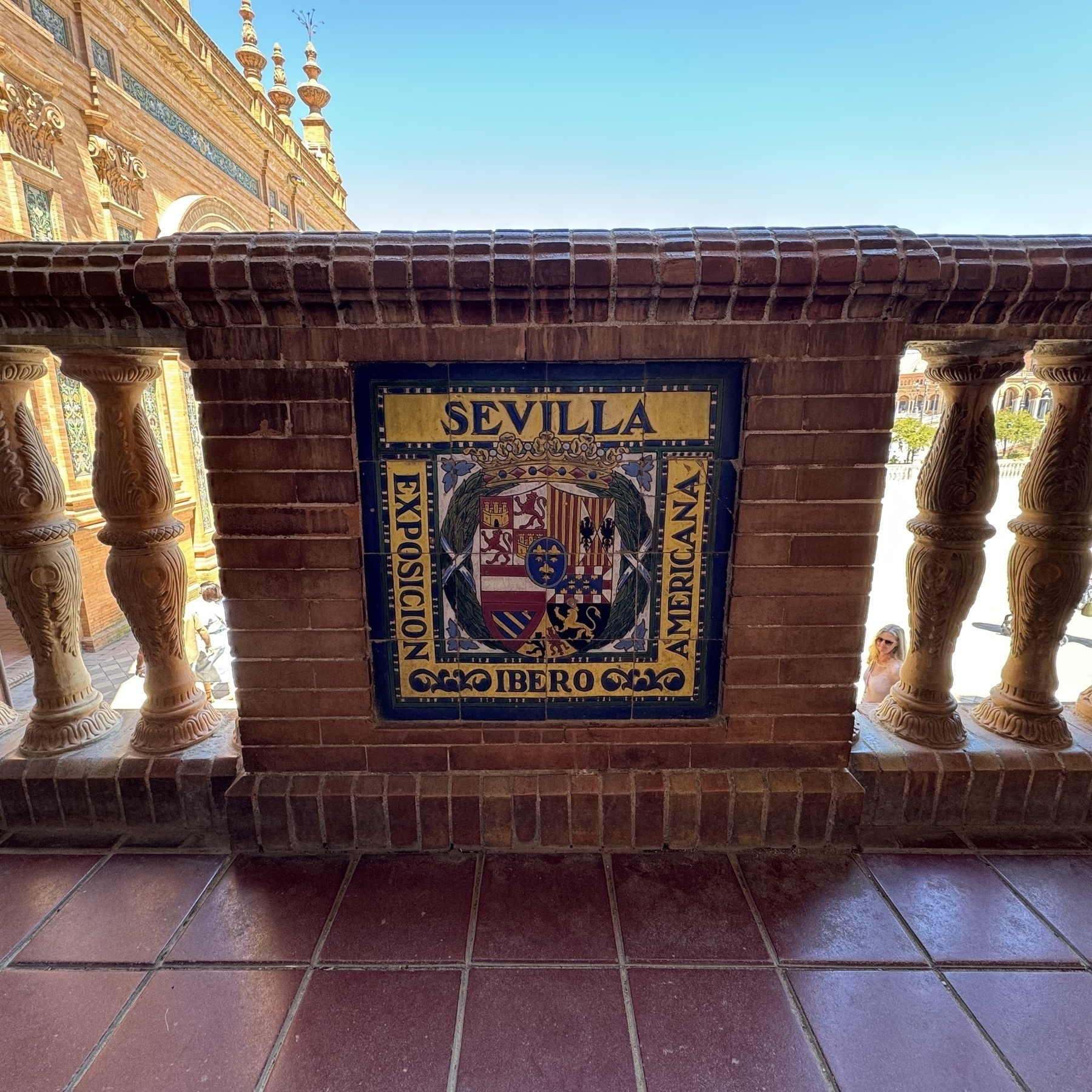 A decorative tile plaque set in a red brick balustrade displays the word “Sevilla” and the crest of the 1929 Ibero-American Exposition. Ornate ceramic columns frame the plaque, with architectural details of the Plaza visible through the opening.