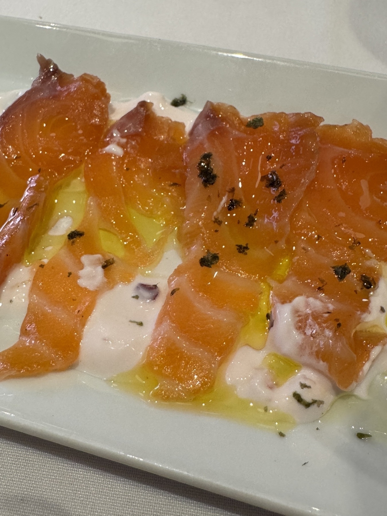 Thin slices of herb-cured salmon laid in elegant rows over a creamy yogurt sauce, finished with a light drizzle of olive oil and scattered herbs that highlight the fish’s marbled texture and vibrant orange hue.
